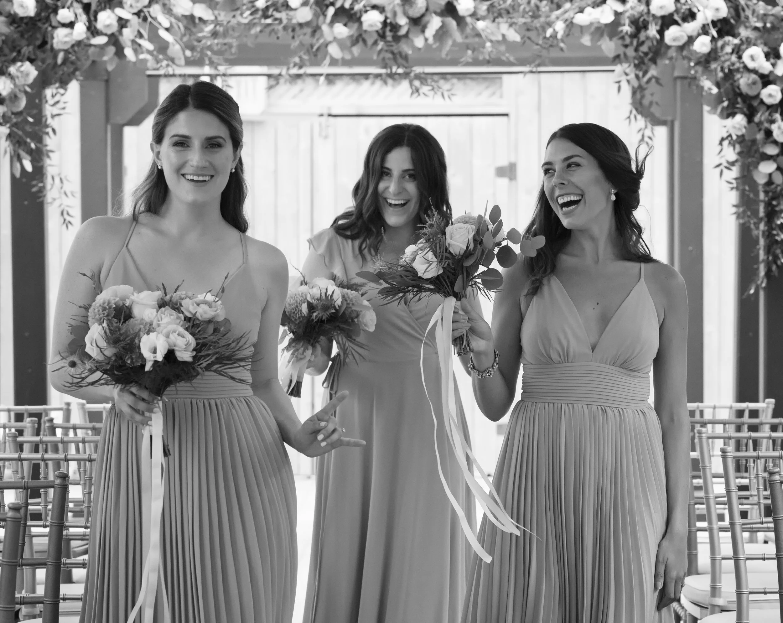 Three women wearing dresses holding bouquets of flowers, smiling and walking in a decorated outdoor venue.