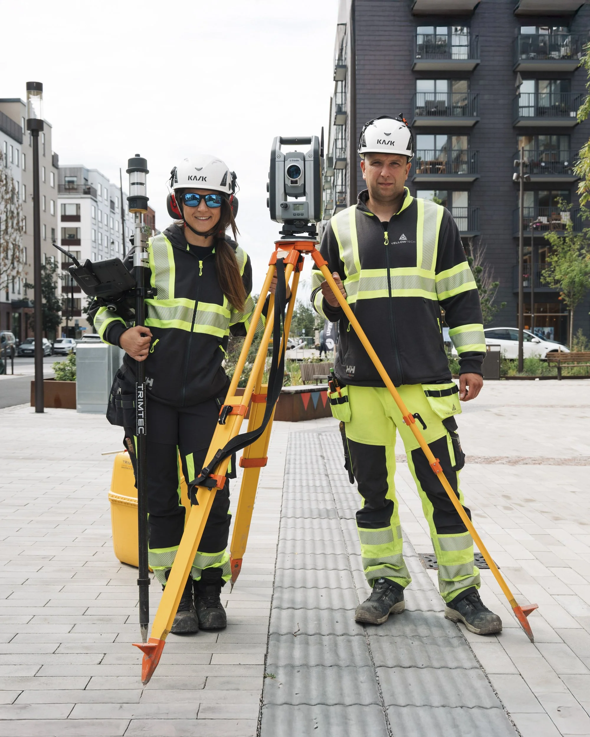 Two construction workers, one woman and one man, standing outdoors on a partially finished paved street with a building in the background. They are wearing safety helmets and high-visibility clothing, and are operating surveying equipment, including 