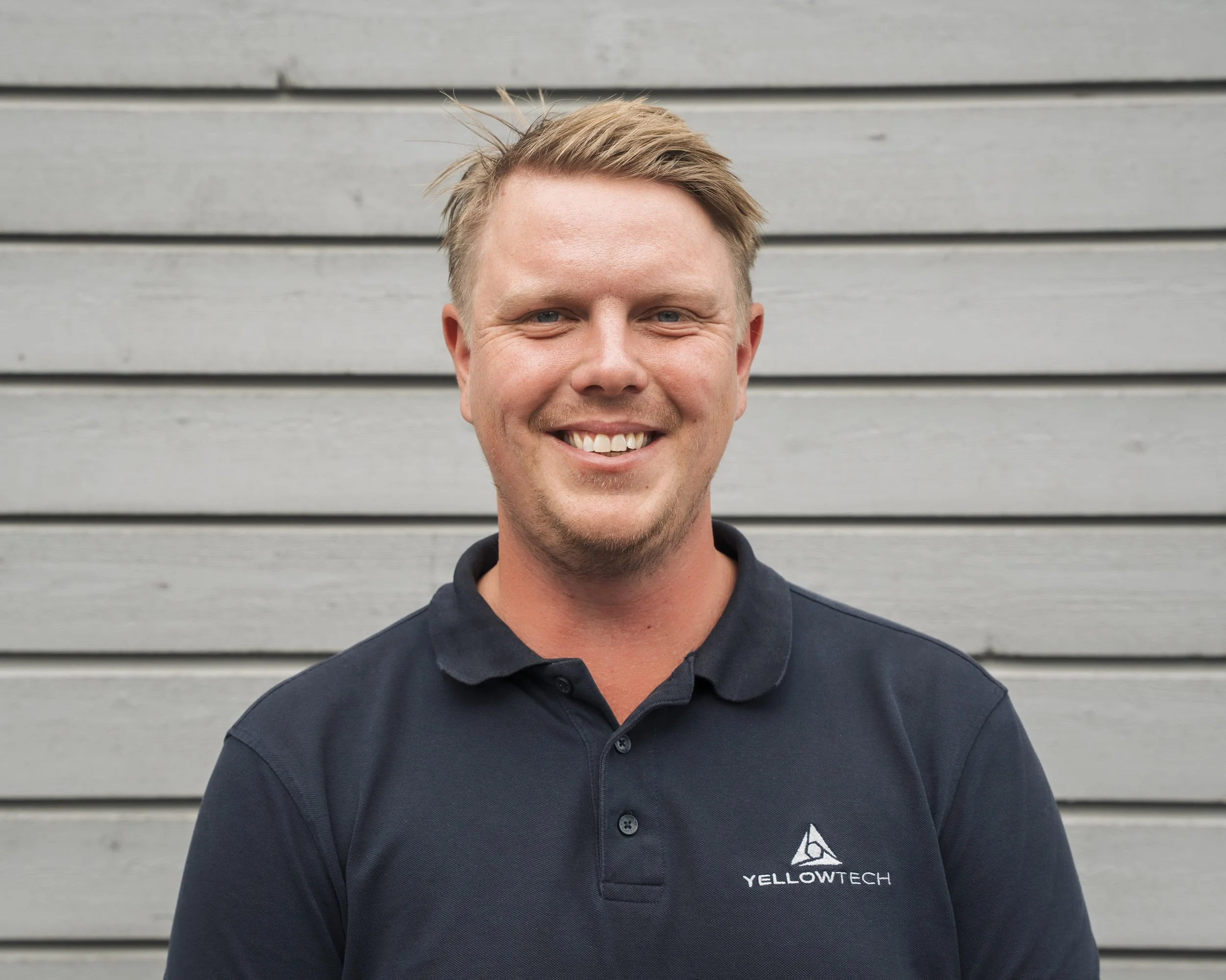 Close-up of a smiling man with short blond hair wearing a dark-colored shirt with a logo, standing in front of a gray wooden wall.