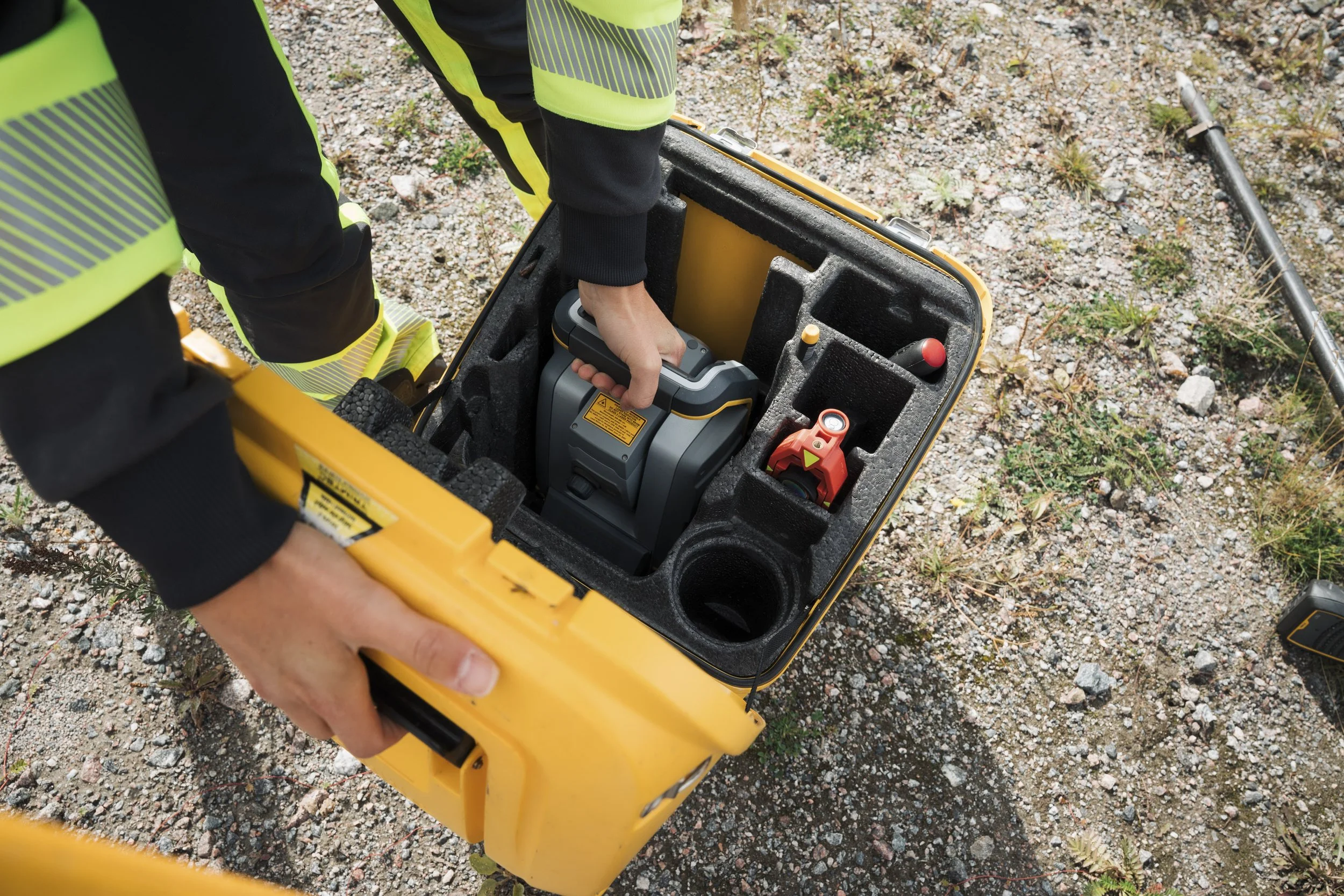 A person wearing a high-visibility safety vest is handling a portable device inside a yellow equipment case on rocky outdoor terrain with sparse vegetation.