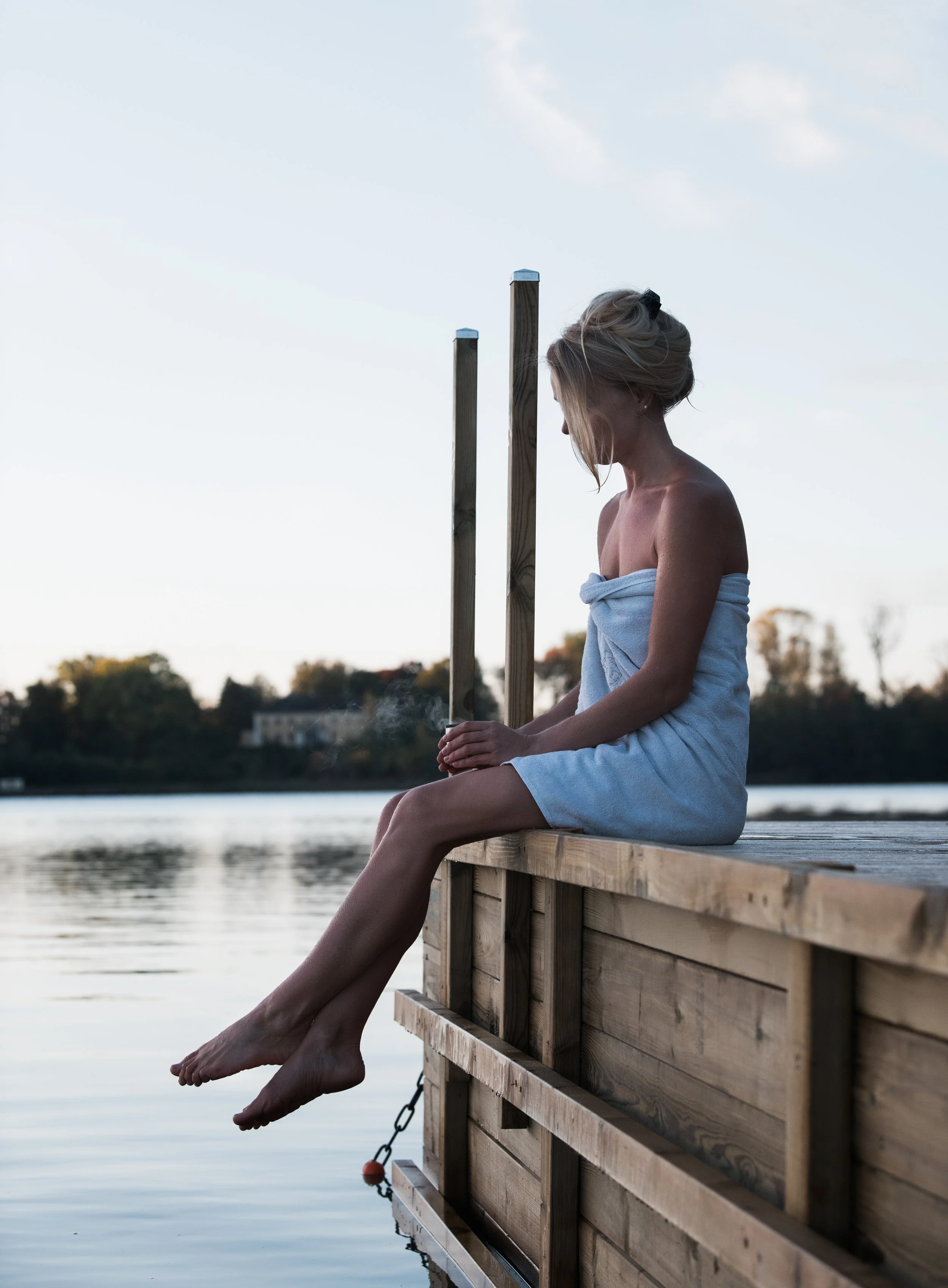 A woman wrapped in a towel sitting on a wooden dock by the water with her legs hanging over the edge, holding two wooden poles, during dusk or dawn.