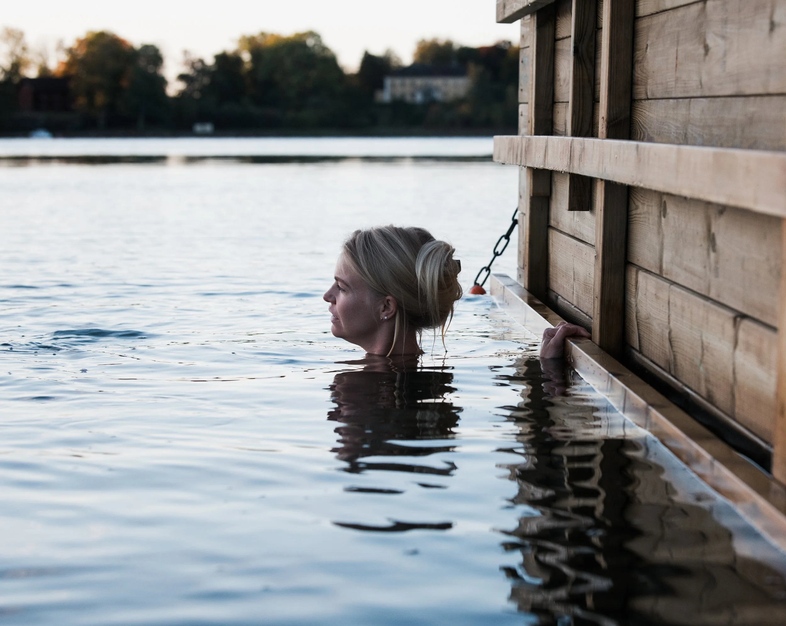 A woman with blonde hair in a bun swimming near a wooden dock by a lake, with trees and houses in the background during sunset.
