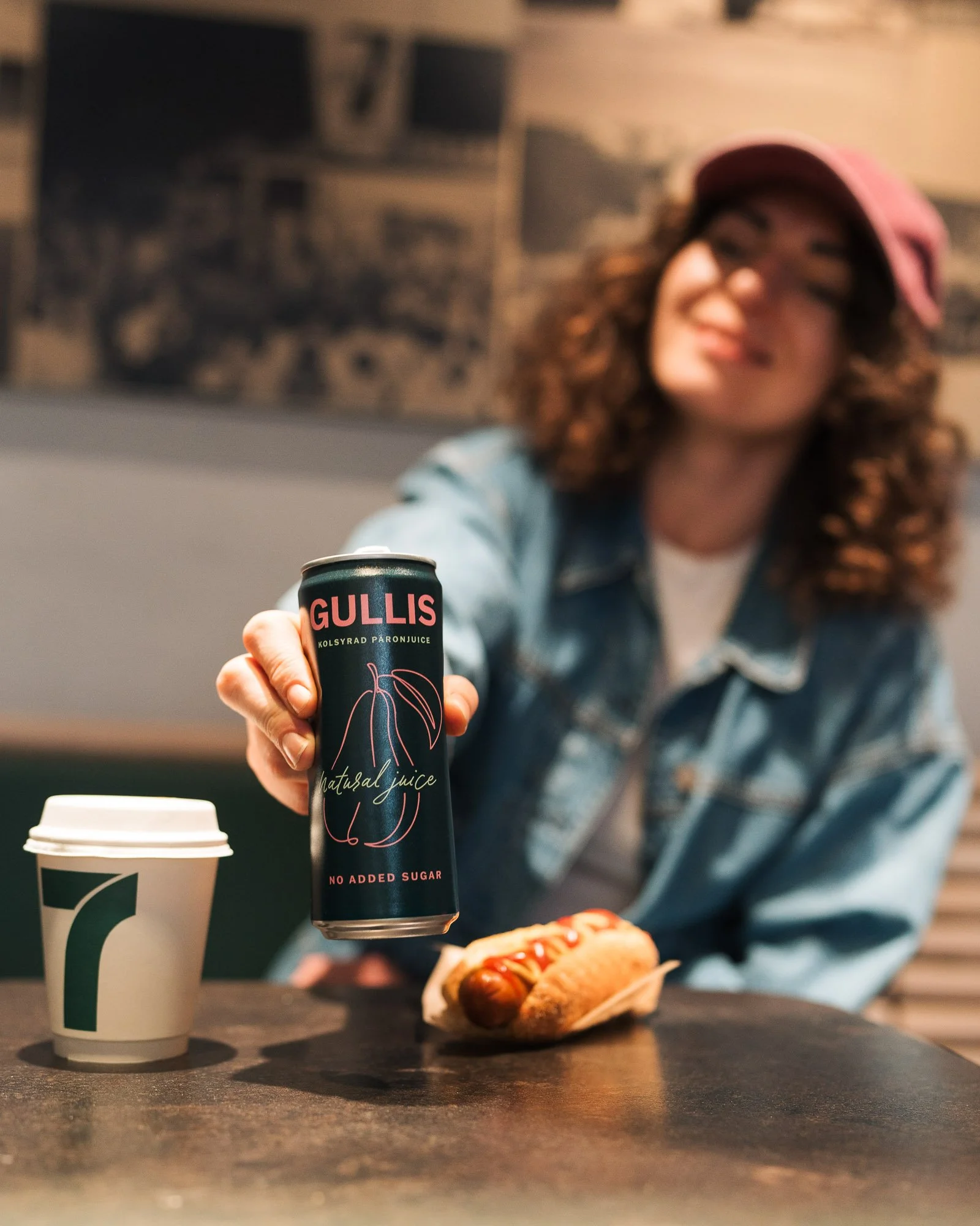A woman in a pink cap and denim jacket is reaching out with a can of Gullis natural juice, with a hot dog and coffee cup on a table in front of her.