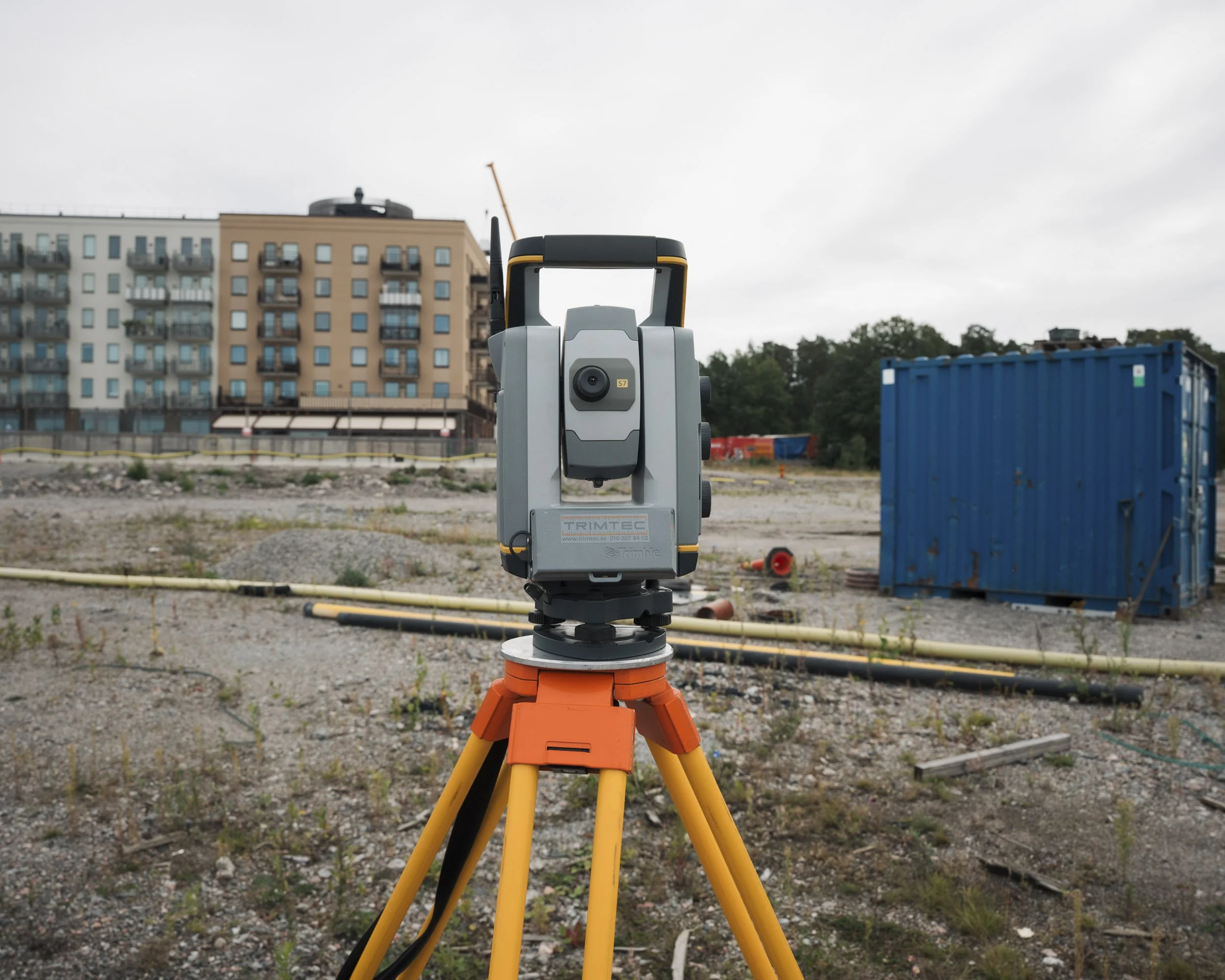 Surveying instrument on tripod at construction site with a building, trees, and portable toilet in the background.