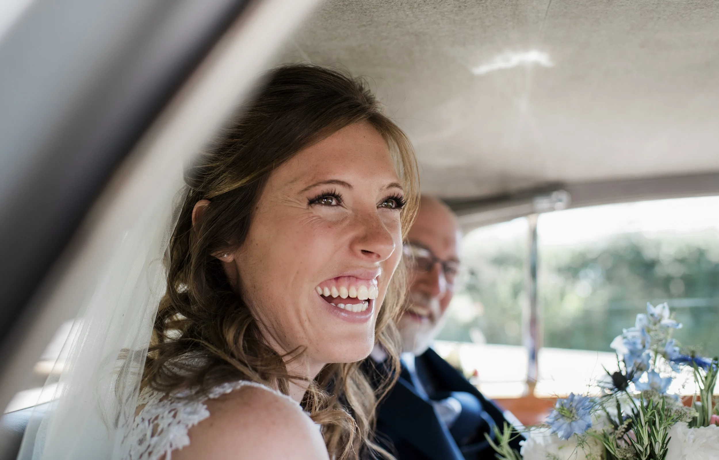 A woman with brown hair and a white dress smiling on a bus or vehicle with a man in a suit next to her, holding flowers.