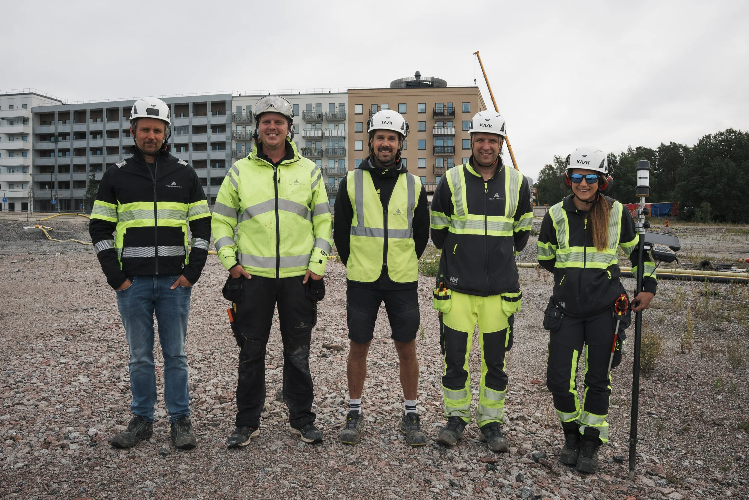 Five construction workers in safety gear and helmets standing on a construction site with apartment buildings in the background.