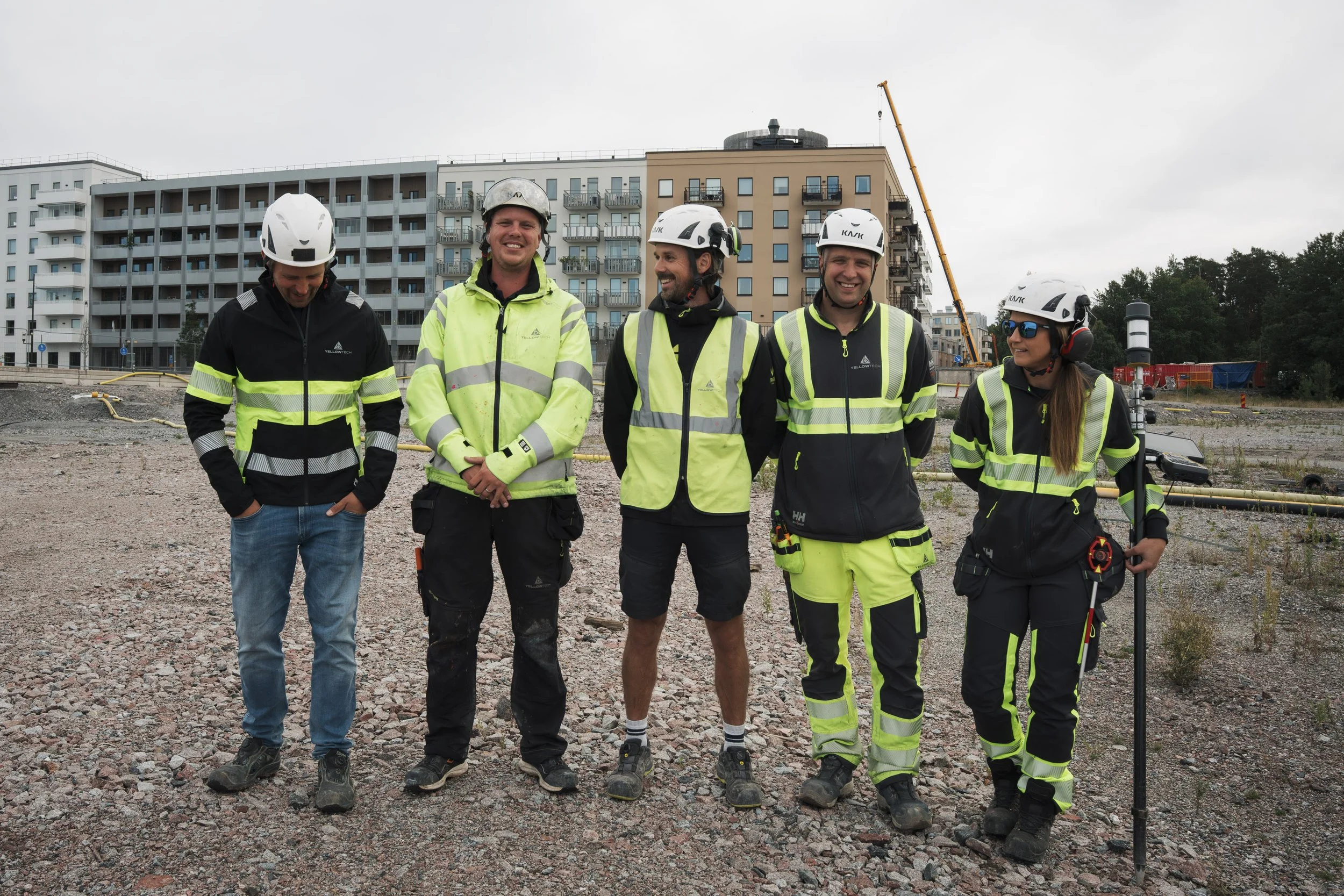 A group of five construction workers, four men and one woman, standing on a gravel construction site with buildings and a crane in the background. They are wearing safety helmets, reflective vests, and work boots, and are smiling and talking.