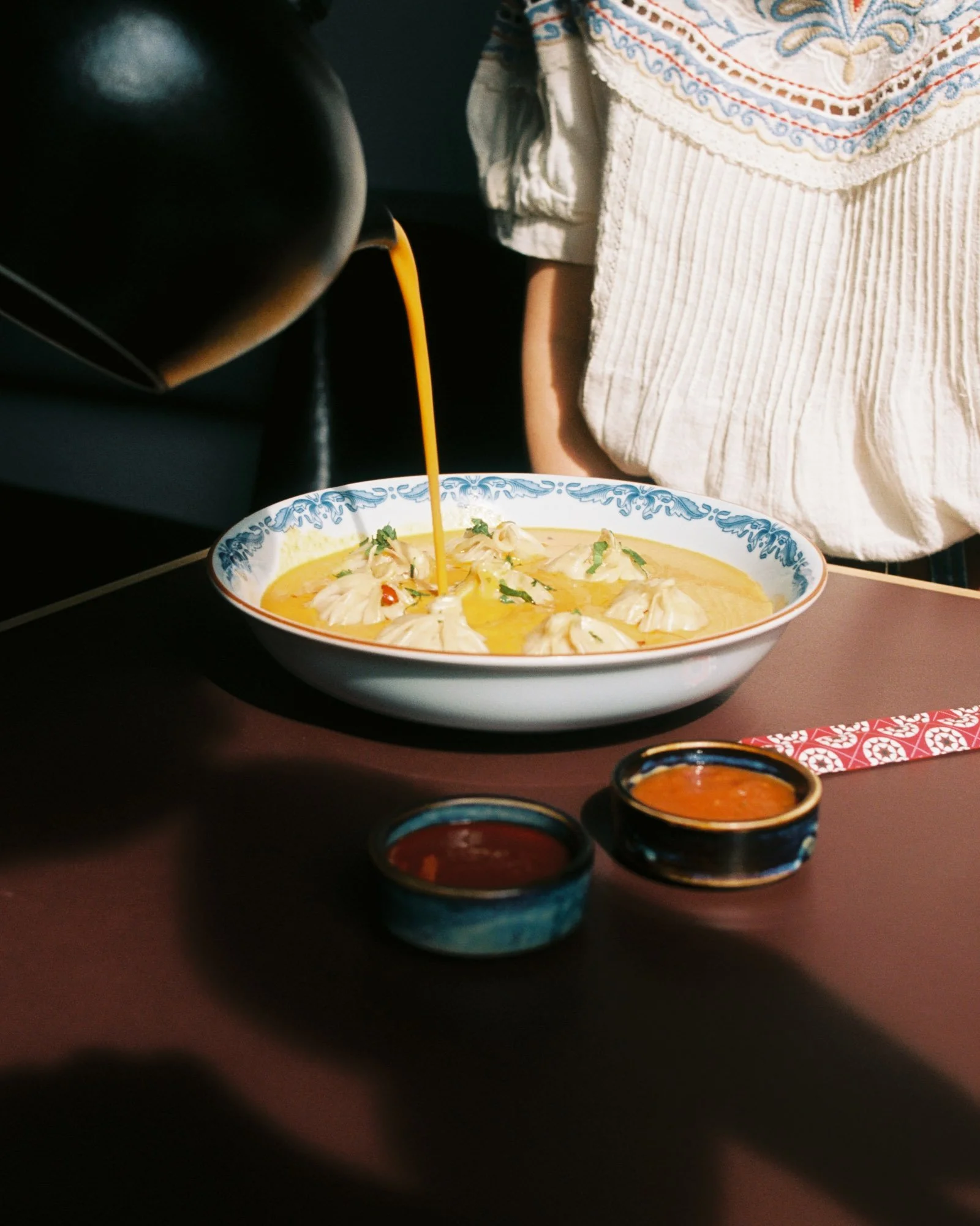 Pour of orange-colored liquid being poured into a bowl with what appears to be soup, with two small bowls of dipping sauces in the foreground, and a person wearing a white embroidered blouse in the background.