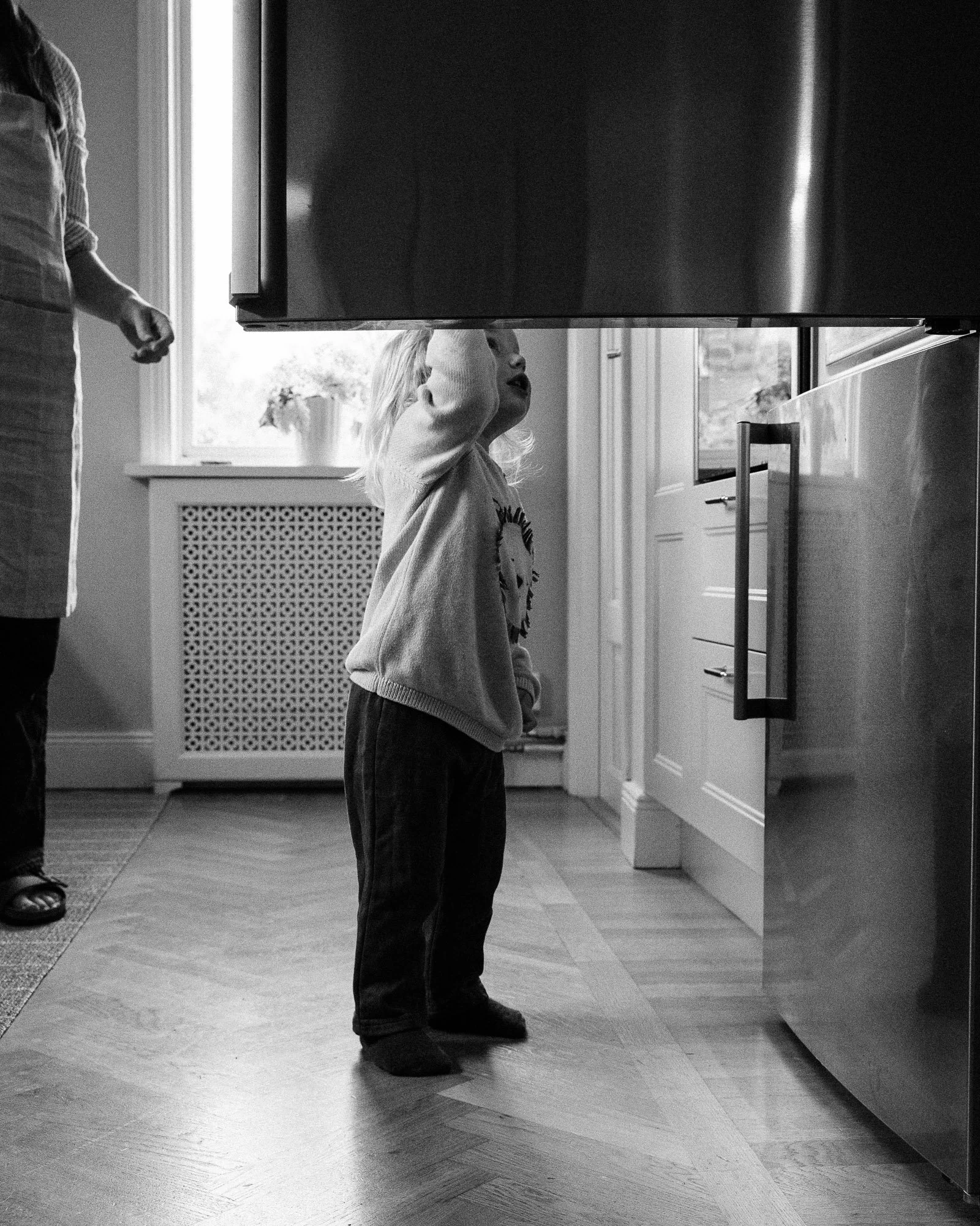A black-and-white photo of a young girl with light hair standing in a kitchen, looking up at a large appliance or object above her. An adult, partially visible on the left, is standing nearby.