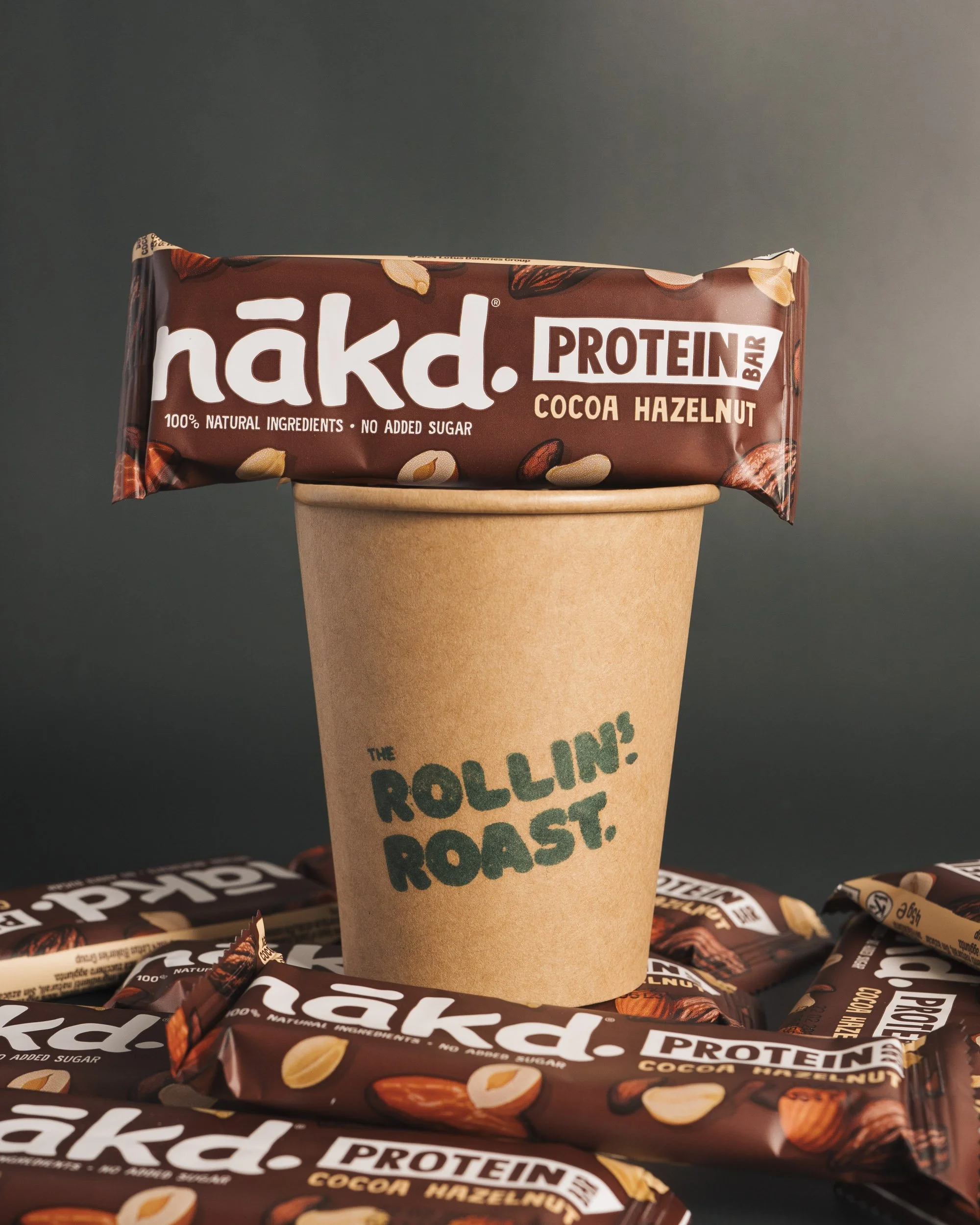 A protein bar in front of a paper cup on a dark background. The protein bar is brown with white text reading 'nakd! PROTEIN BAR' and 'COCOA HAZELNUT'. The paper cup has green text that appears to say 'THE POLLINS?'.