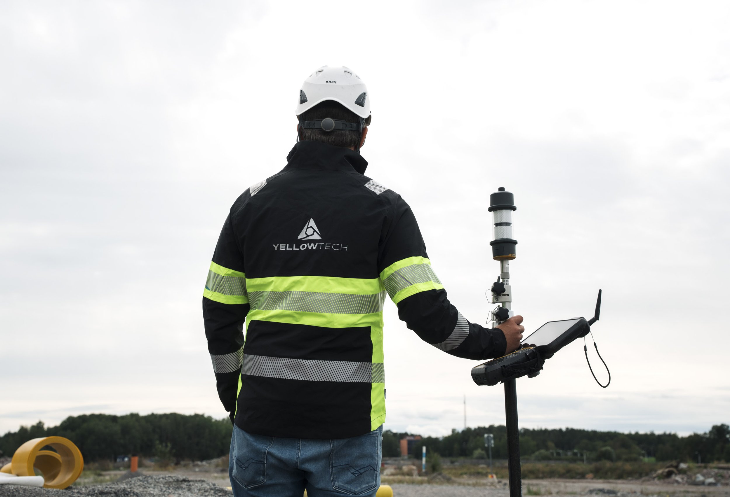 A person wearing a black and yellow high-visibility jacket and a white hard hat standing outdoors, operating surveying equipment on a tripod.