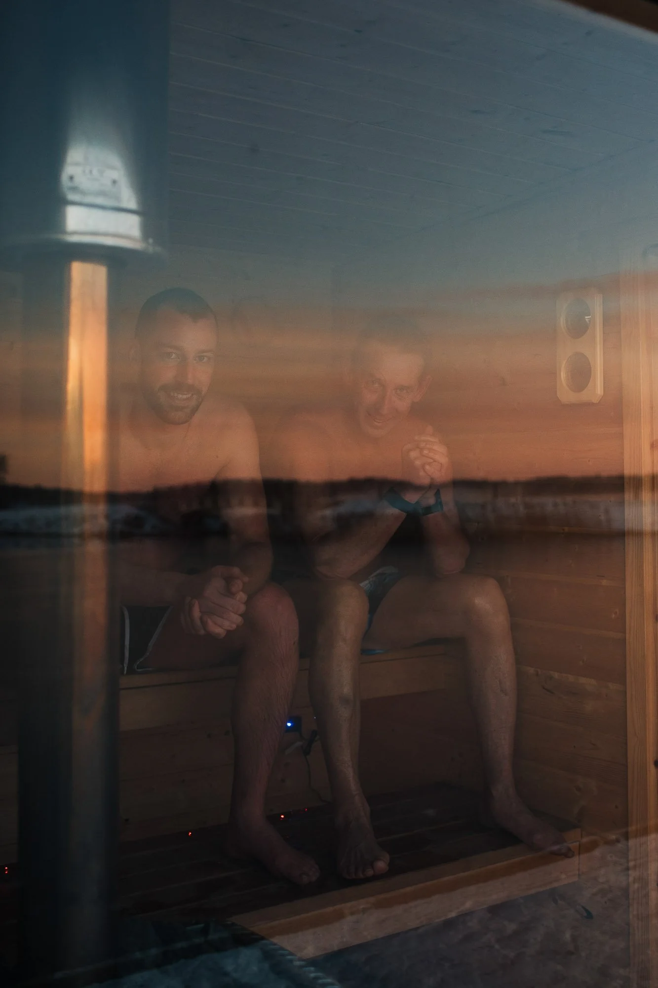 Two men sitting inside a wooden sauna, smiling, with steam visible on the glass door, and a wall-mounted speaker on the wall.