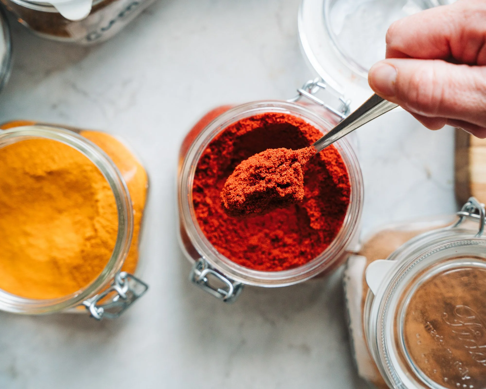 A close-up of a hand scooping red chili powder from a glass jar with a metal spoon. Several other spice jars with red, orange, and brown spices are visible around it.