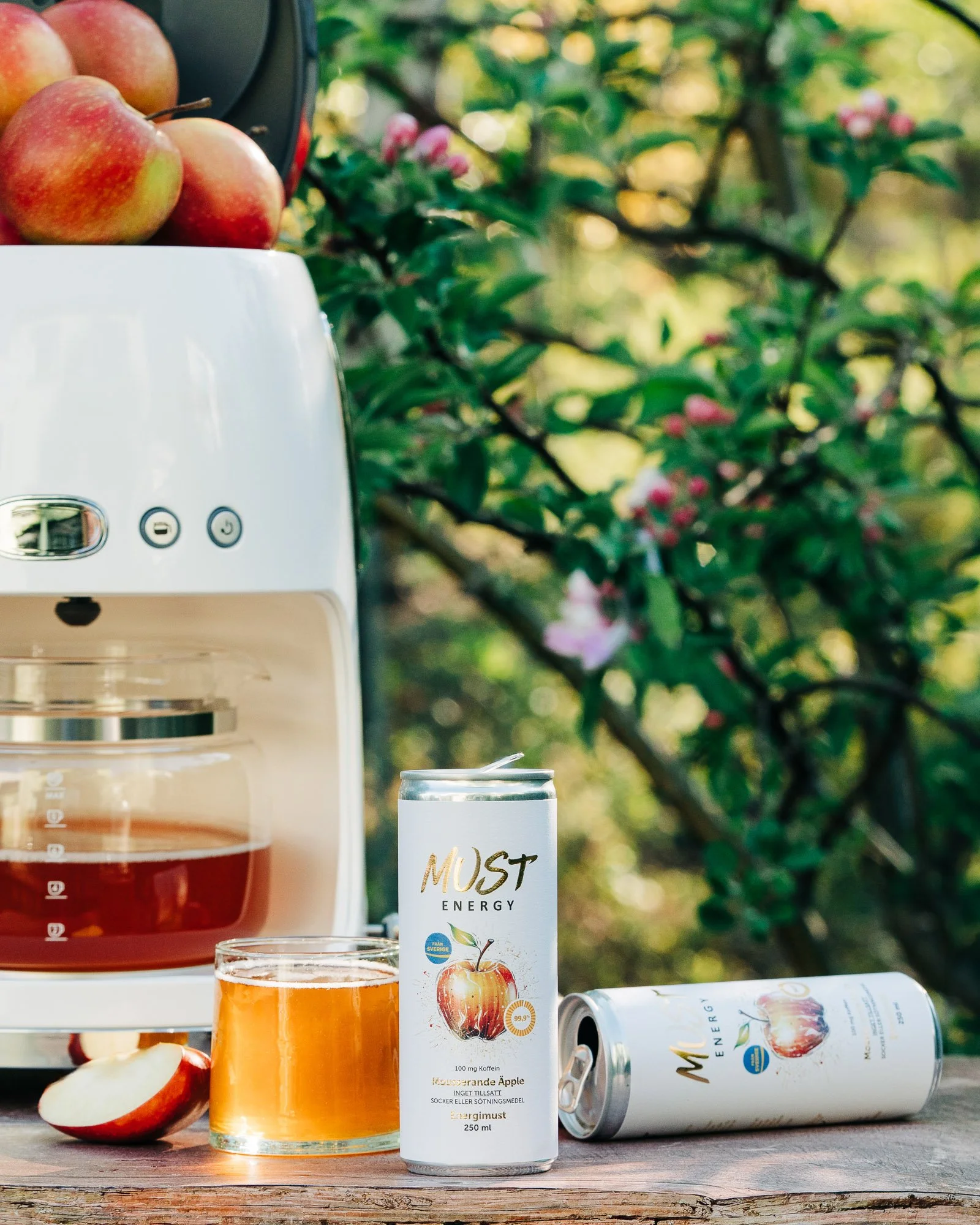A white energy drink can labeled 'MUST ENERGY' with an apple illustration, an open glass of orange-colored beverage, a white coffee machine, and a sliced apple on a wooden surface outdoors with green foliage in the background.