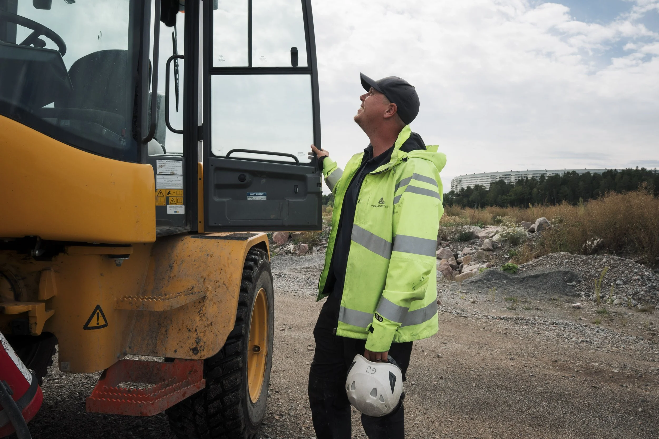 A construction worker wearing a yellow safety jacket and black cap, holding a white safety helmet and a white helmet on the ground, standing next to a yellow construction vehicle outdoors with a cloudy sky and trees in the background.