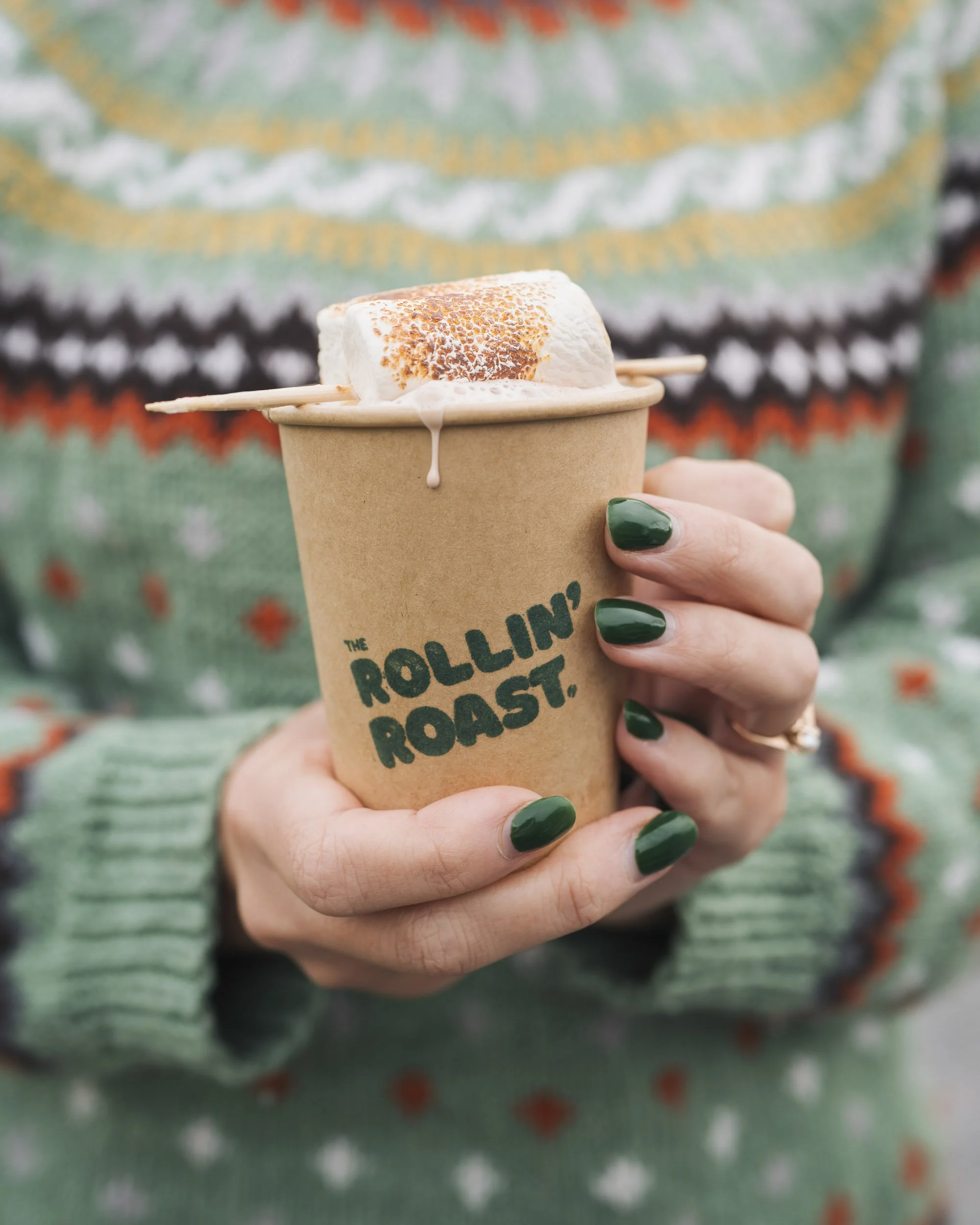 Person holding a cup of roasted marshmallow on a stick, with a colorful knit sweater in the background.
