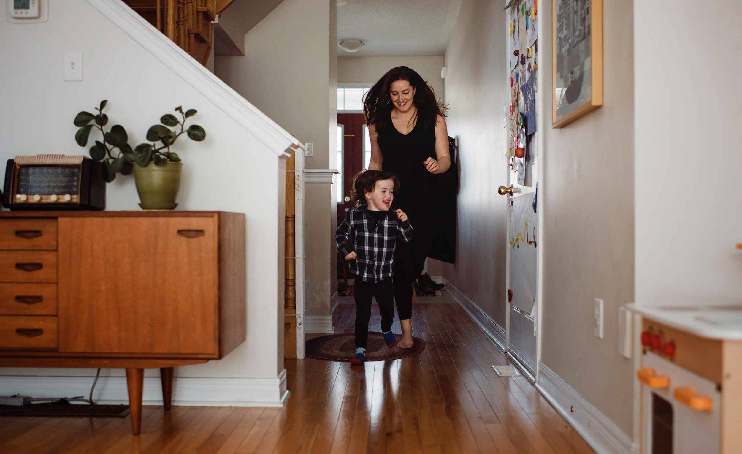 A woman and a little girl walking down a hallway inside a home, smiling and looking at each other.