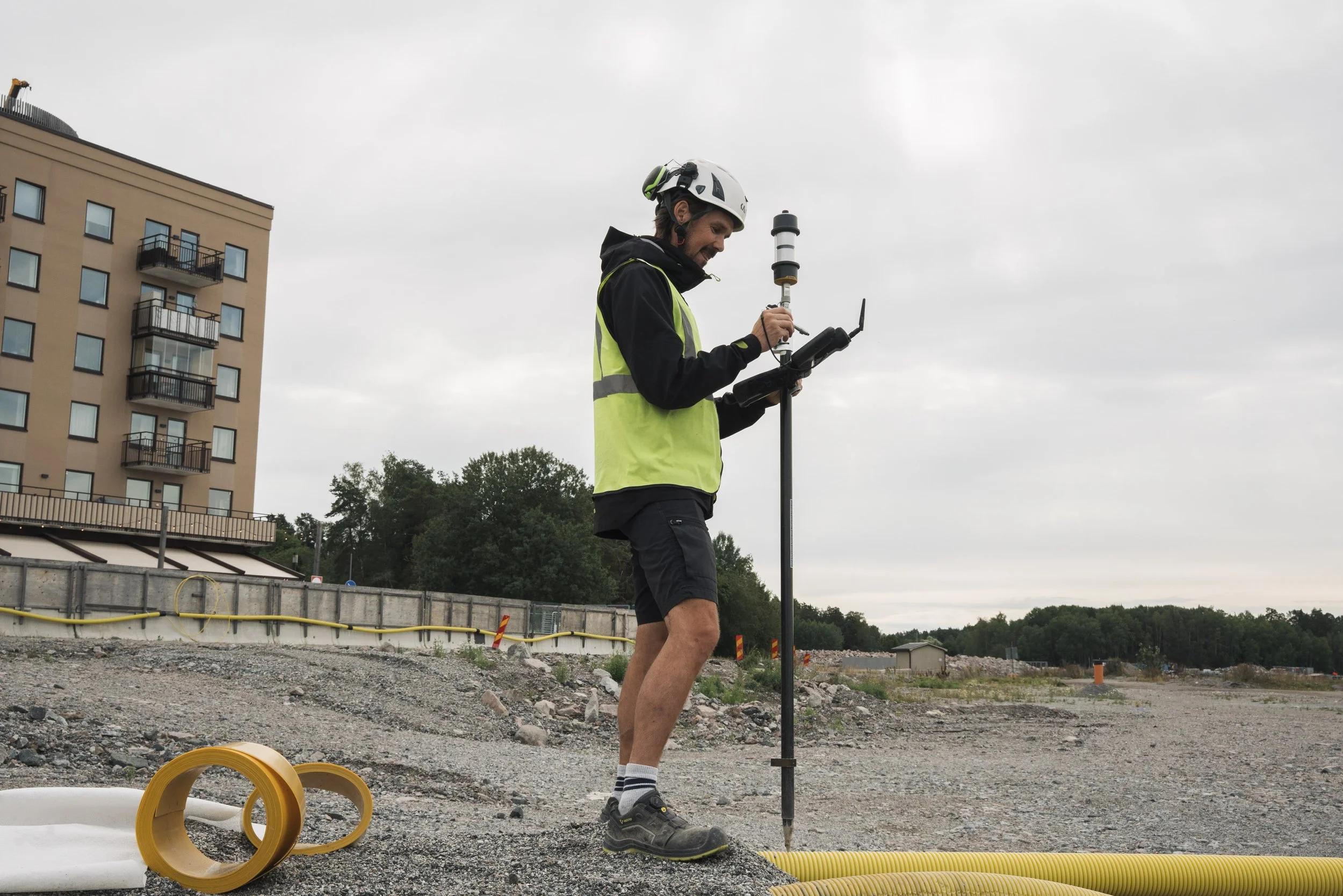 A man wearing a white helmet, black jacket, yellow safety vest, and shorts is standing on an outdoor construction site. He is holding a ladder and a tablet, surrounded by yellow and white construction materials and pipes, with a building and trees in