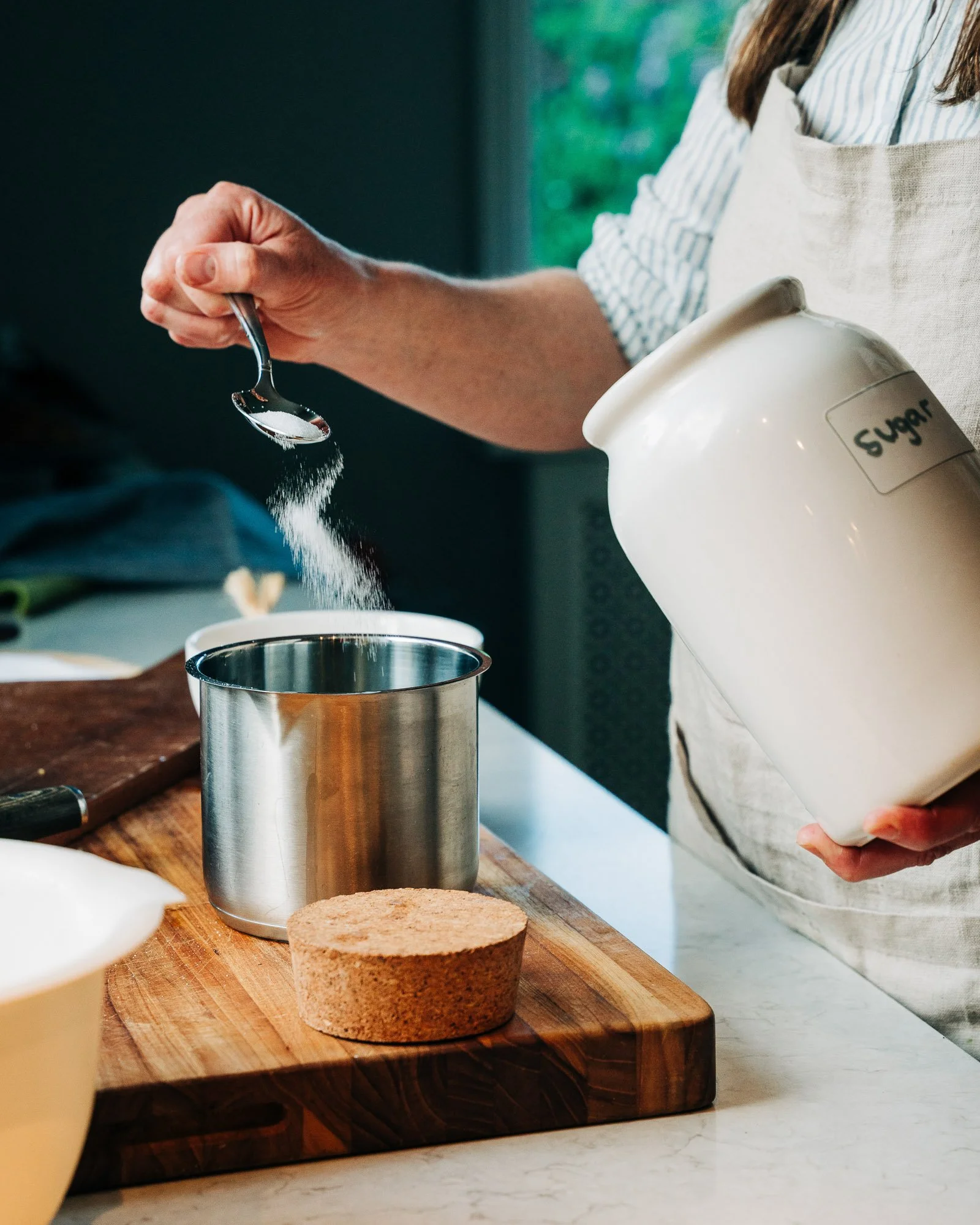 A person holding a spoon with sugar over a container labeled 'sugar'. The person is wearing a striped shirt and apron, standing at a kitchen counter with a wooden cutting board, a cork container, and a white bowl.