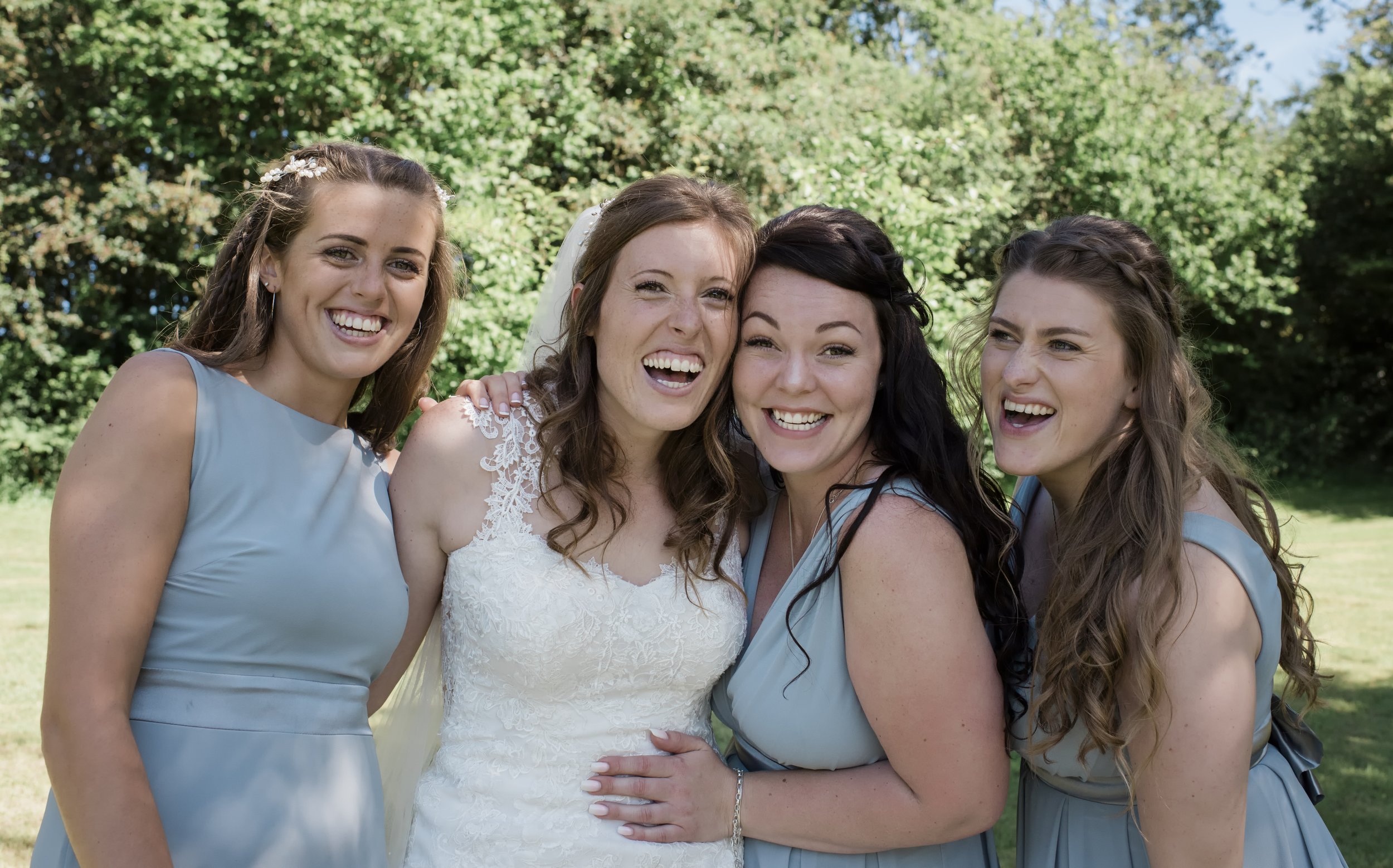 A group of four women smiling and posing outdoors on a sunny day, with three of them wearing matching blue dresses and one in a white lace dress, suggesting a wedding celebration.