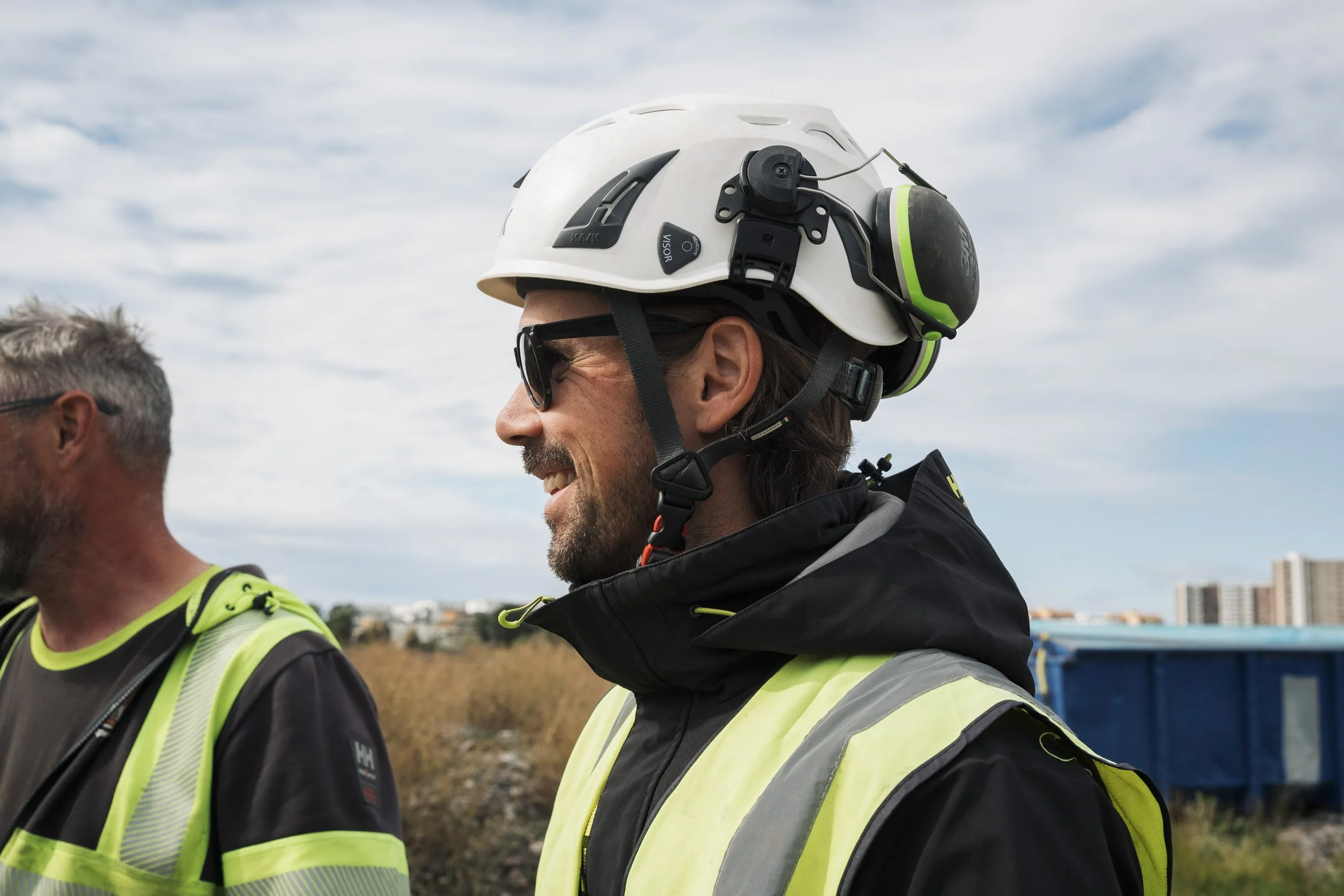 Two men wearing sunglasses and safety vests stand outdoors under a partly cloudy sky. One man is smiling and wearing a white helmet with mounted ear protection, the other man’s face is partially visible.