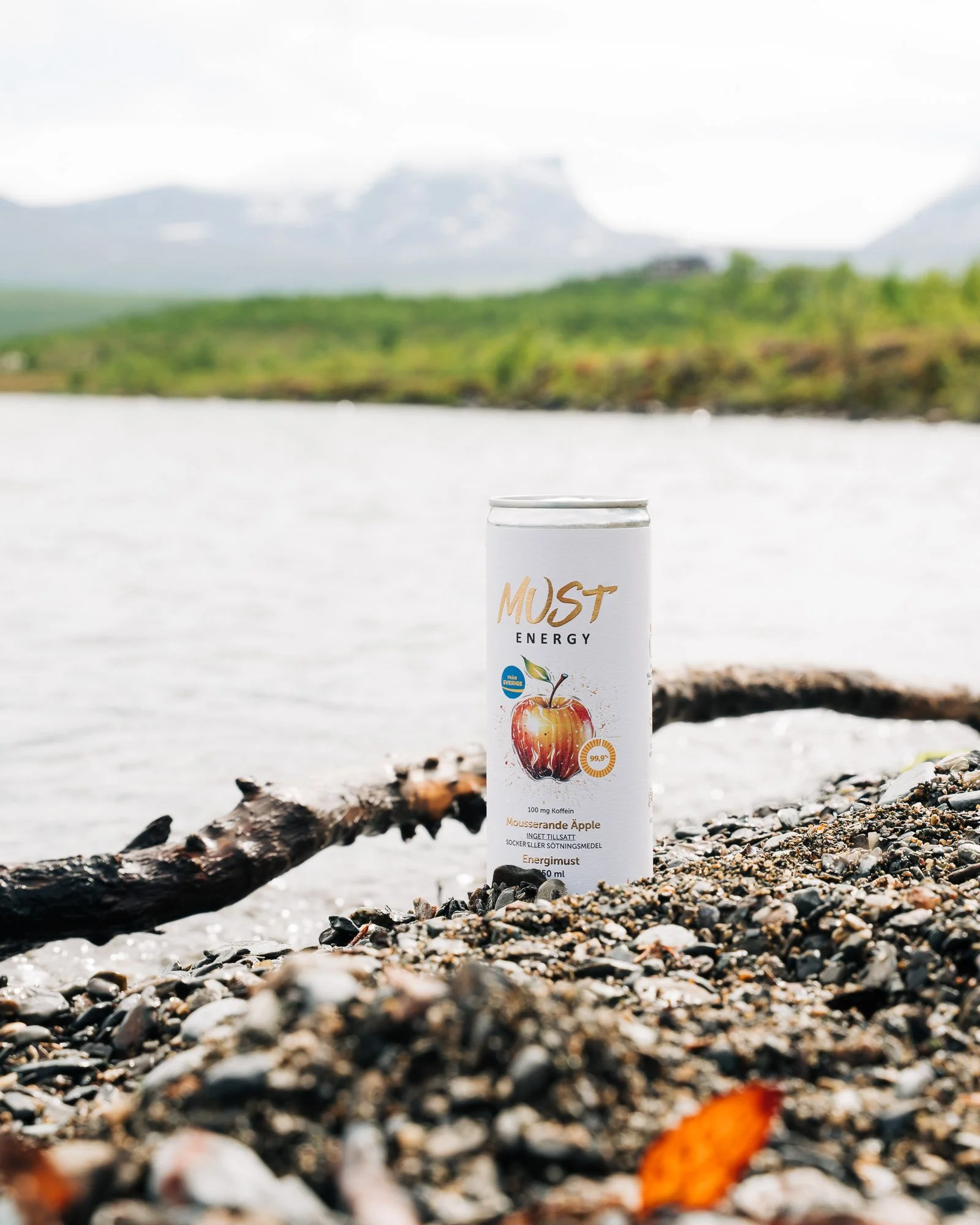 A white can of energy drink labeled "MUST ENERGY" with an apple graphic, placed on a rocky beach with a blurred natural landscape and water in the background.
