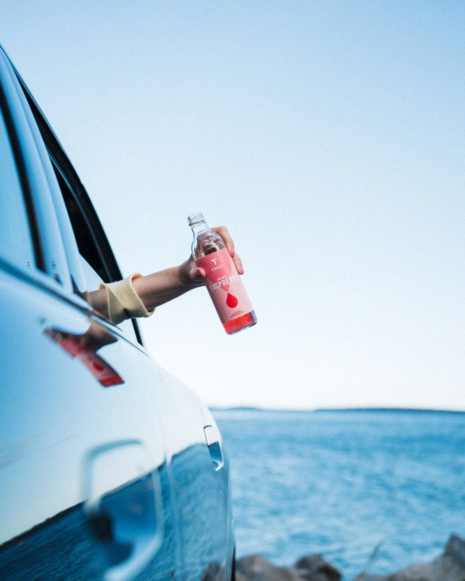 A person's hand holding a bottle of organic raspberry sparkling water, leaning out of a car window with a view of the ocean and a rocky shore in the background.