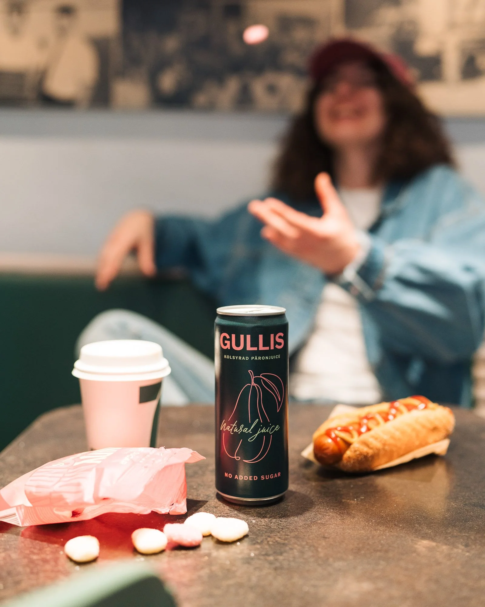 A can of Gullis natural juice with a drawing of a peach and the text "no added sugar" on a table, alongside a hot dog with ketchup and mustard, a paper coffee cup, and some scattered candy
