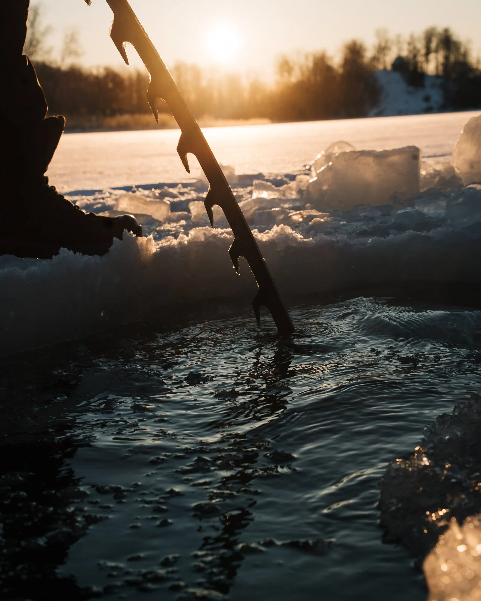 Close-up of an ice fishing scene during sunset with a person holding an ice auger, ice chunks, and icy water with a snowy landscape and trees in the background.