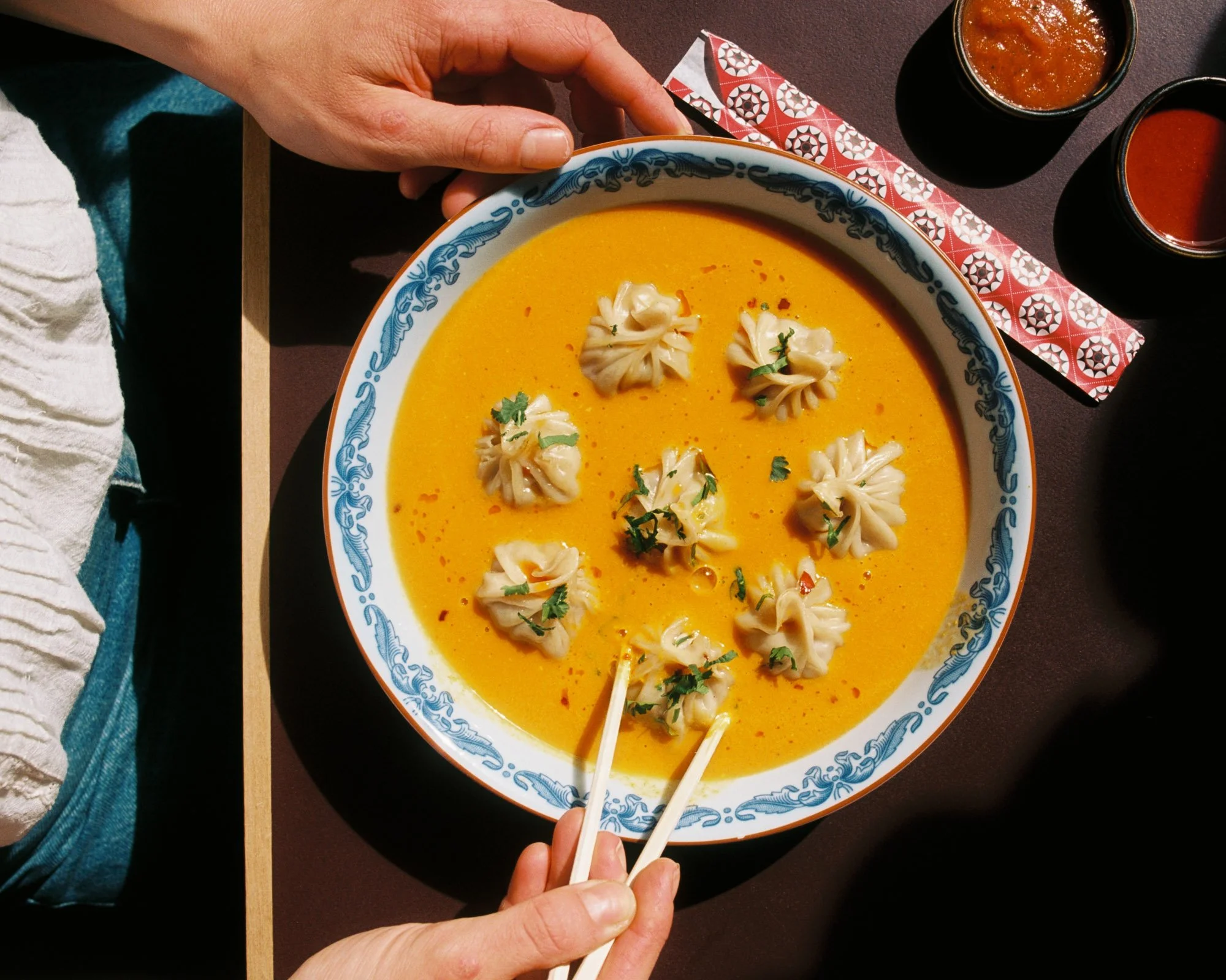 A person using chopsticks to pick up a dumpling from a bowl of orange-colored soup or curry garnished with herbs. Two other dumplings are floating in the soup, and three small bowls of red sauces are nearby on a dark table.