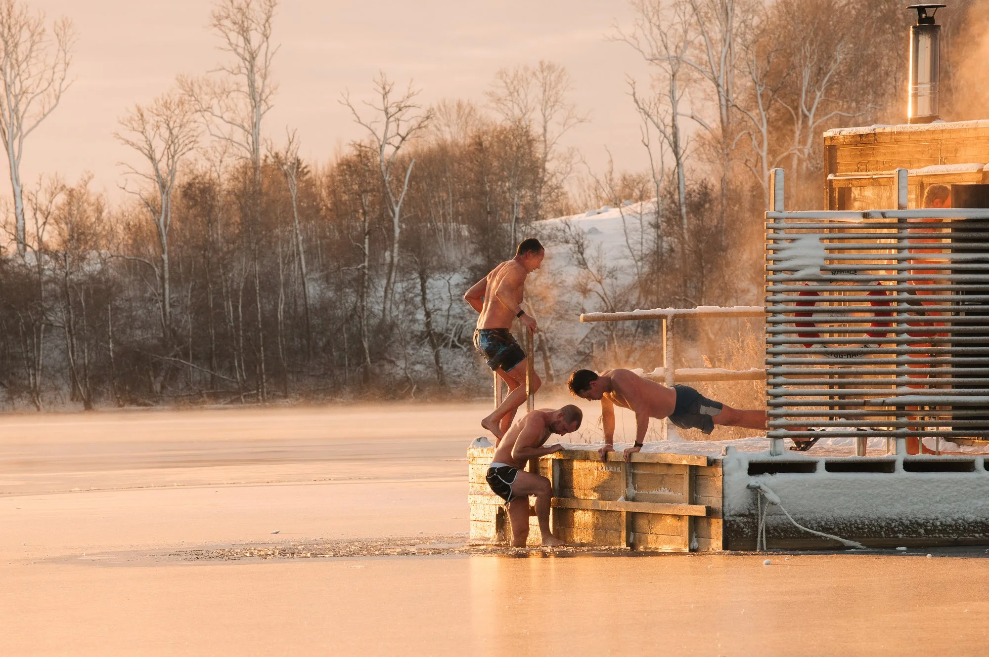 Four people in swimwear, likely participating in a winter ice bath or swimming activity, just outside a heated cabin on a snowy, icy landscape during sunset or sunrise.
