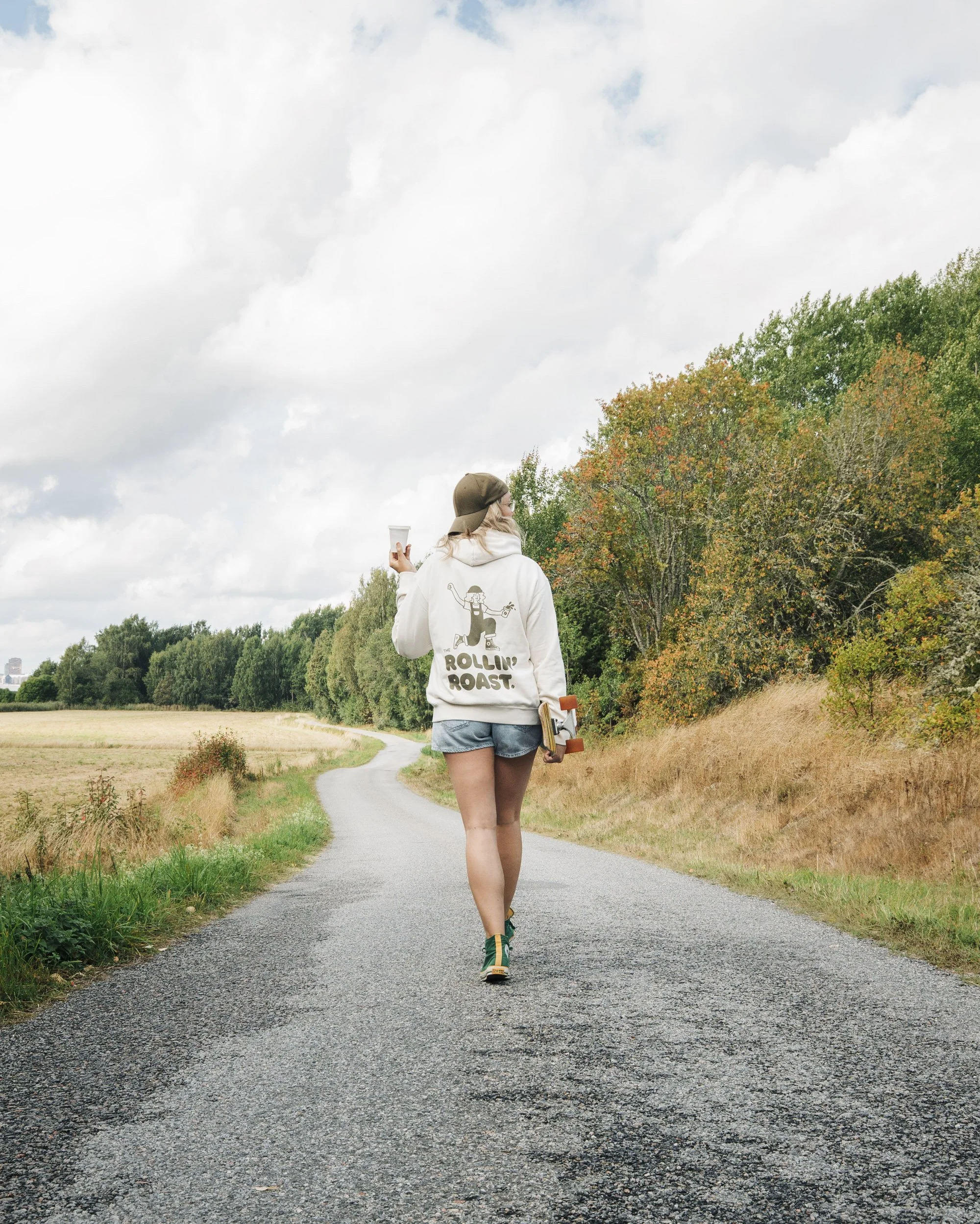 A woman with long hair, wearing a tan cap and a white hoodie with a cartoon figure and text that says 'The Rollin' Roast', is walking on a curved paved path through a grassy field with trees showing autumn leaves, holding a cup in her right hand and 