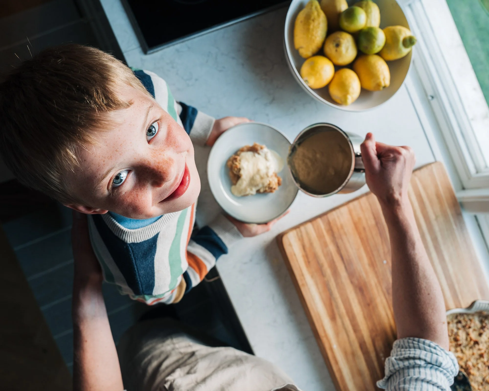 A young boy with red hair and blue eyes looking up at the camera while holding a mug, standing at a kitchen counter with a plate of food, a bowl of lemons, and a glass of juice nearby.