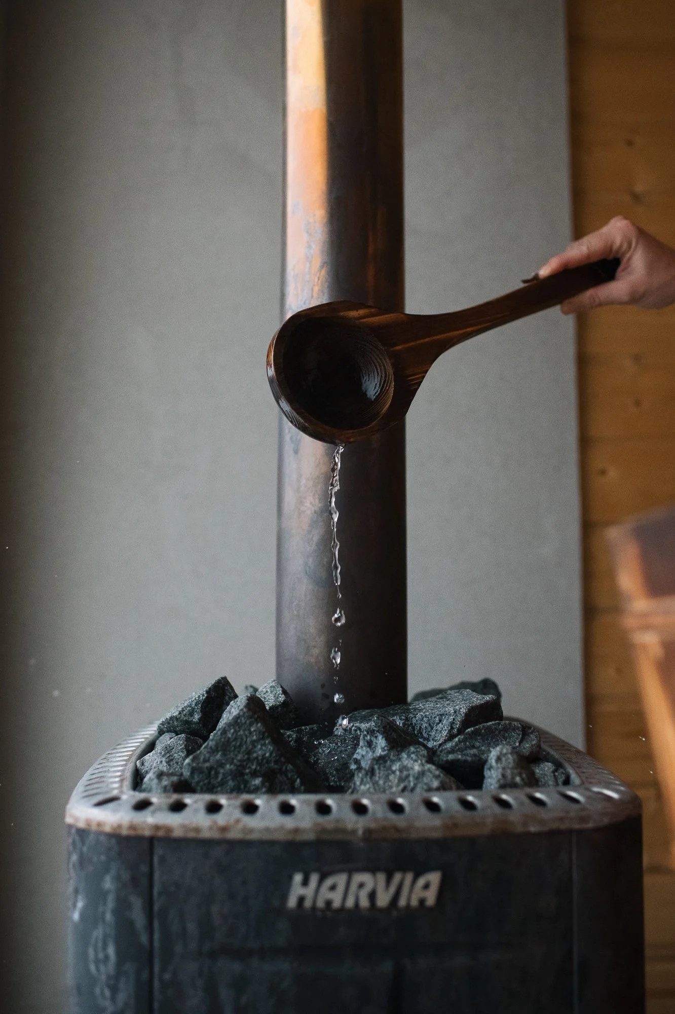 A hand pouring water from a wooden ladle onto hot stones inside a small, rounded, metal container with the word 'HARVIA' on it, with a gray wall and wooden wall in the background.