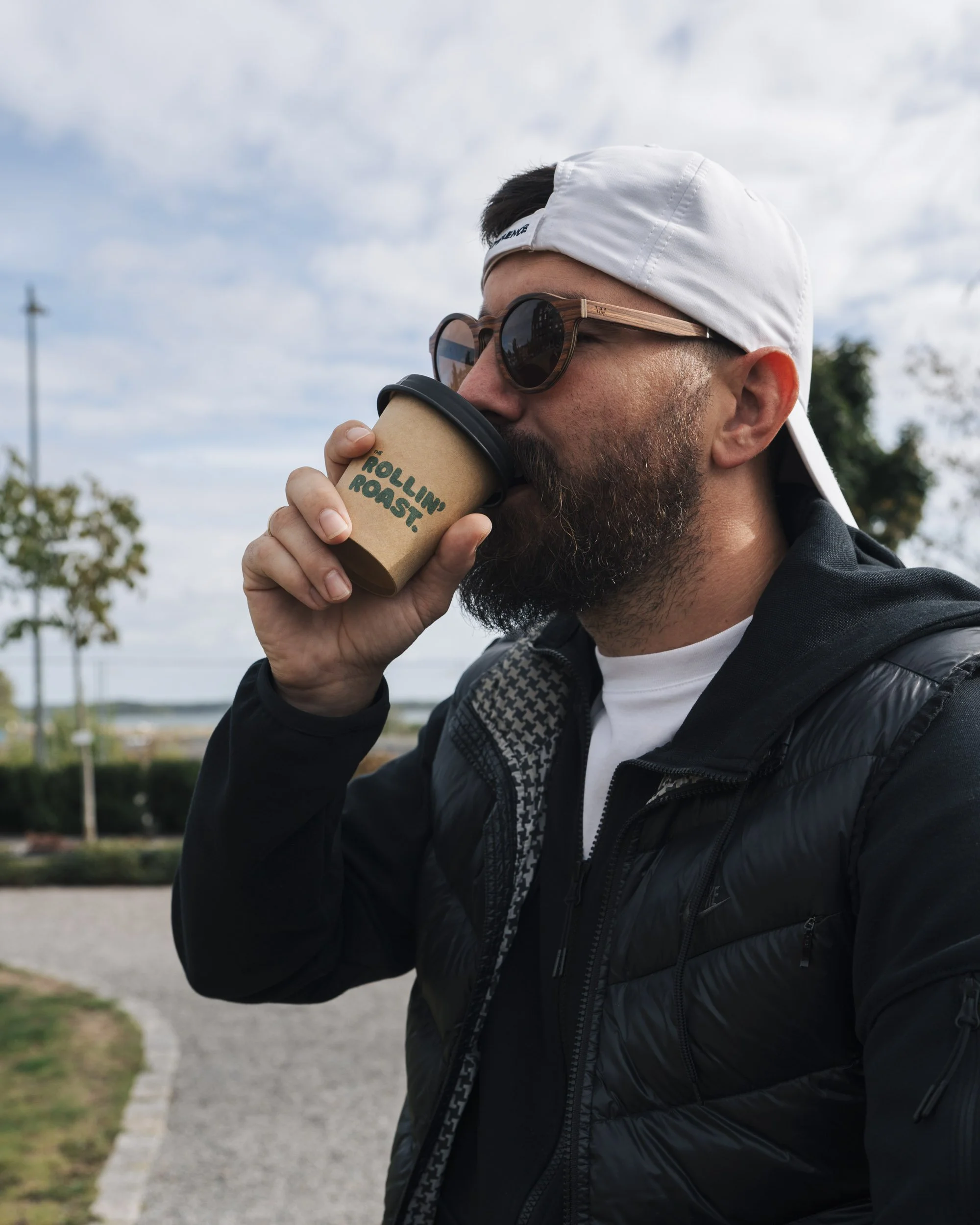 Man with a beard in sunglasses and a backward white cap drinking coffee outdoors, wearing a black jacket, with trees and a cloudy sky in the background.