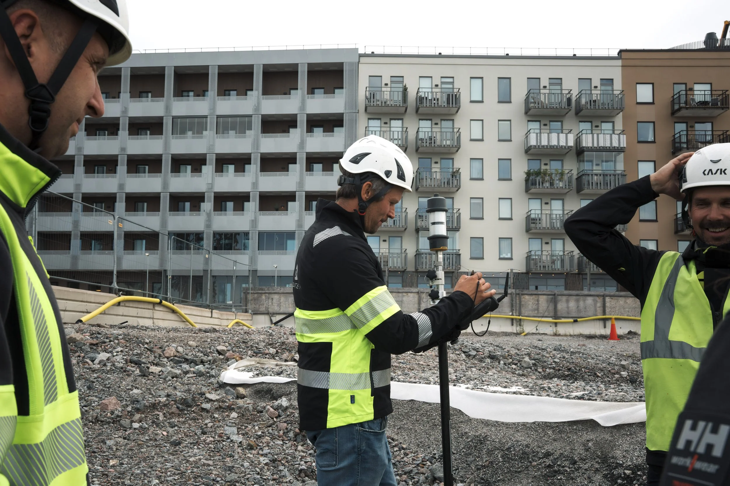 Three construction workers in safety helmets and vests at a construction site with a multi-story residential building in the background, inspecting equipment.