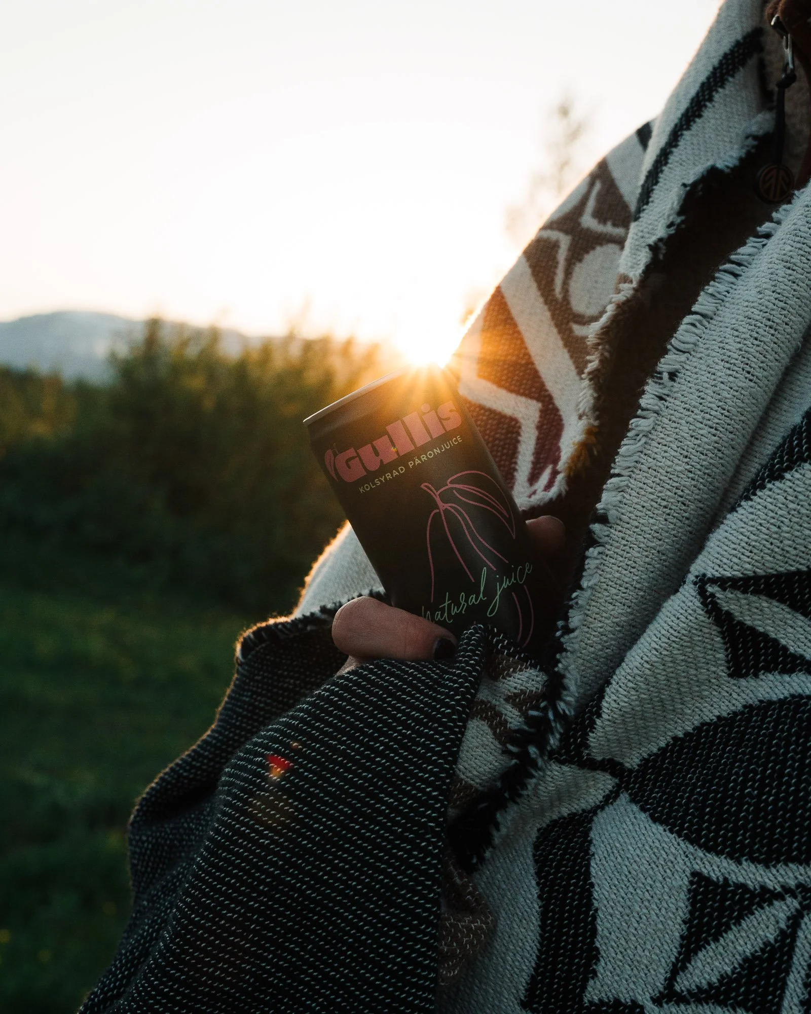 Person holding a cup of juice outdoors during sunset with a blanket wrapped around their shoulders.