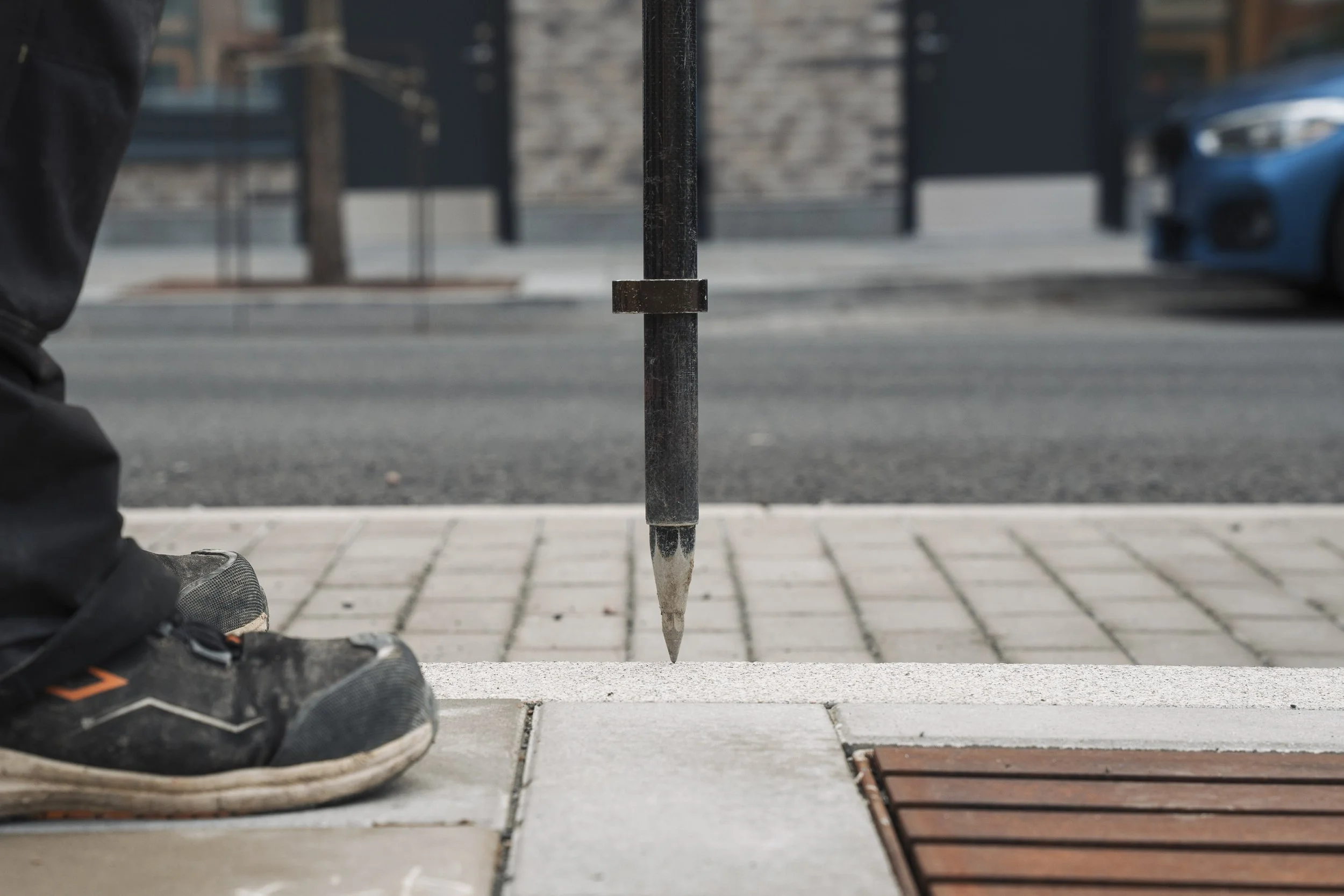 Close-up of a person's foot wearing a sneaker near a metal rod with a pointed end set on a sidewalk, with a building and a parked blue car in the background.