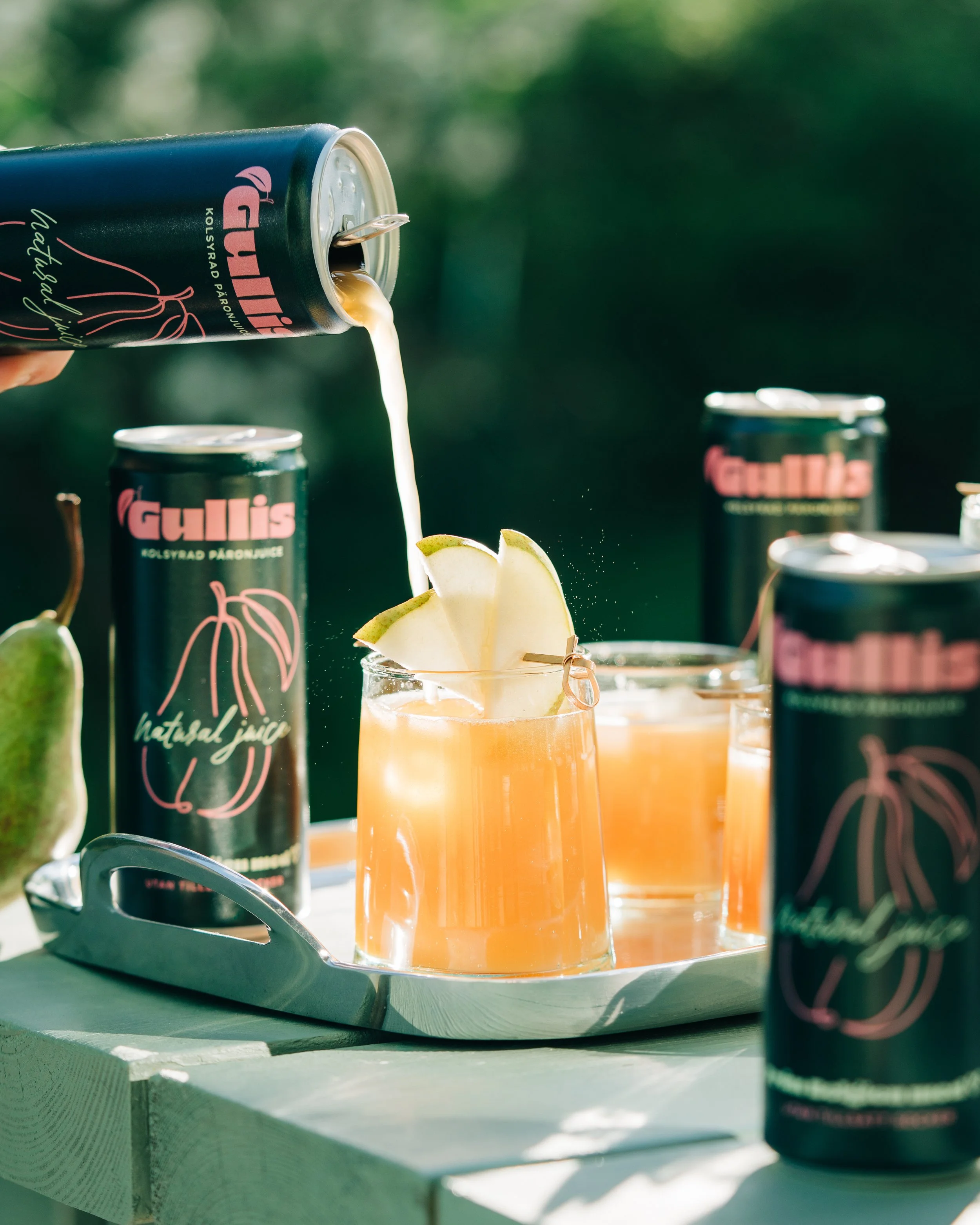 A hand pouring a drink from a can of Gullis into a glass with apple slices, set on a silver tray with other cans and apple slices nearby, outdoors with a blurred green background.
