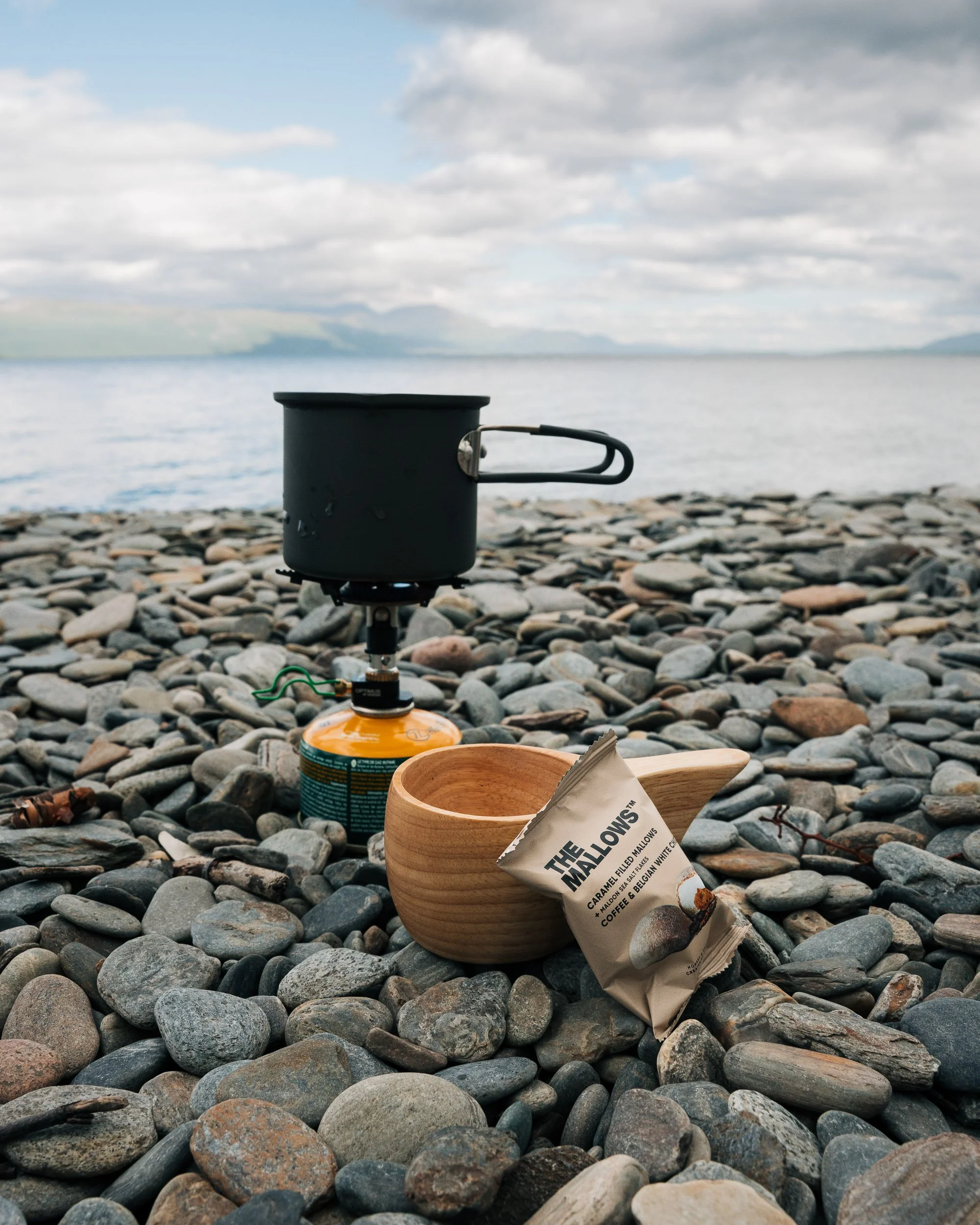 A camping stove with a pot on top sits on a rocky beach with stones and pebbles, overlooking a body of water and a cloudy sky in the background.