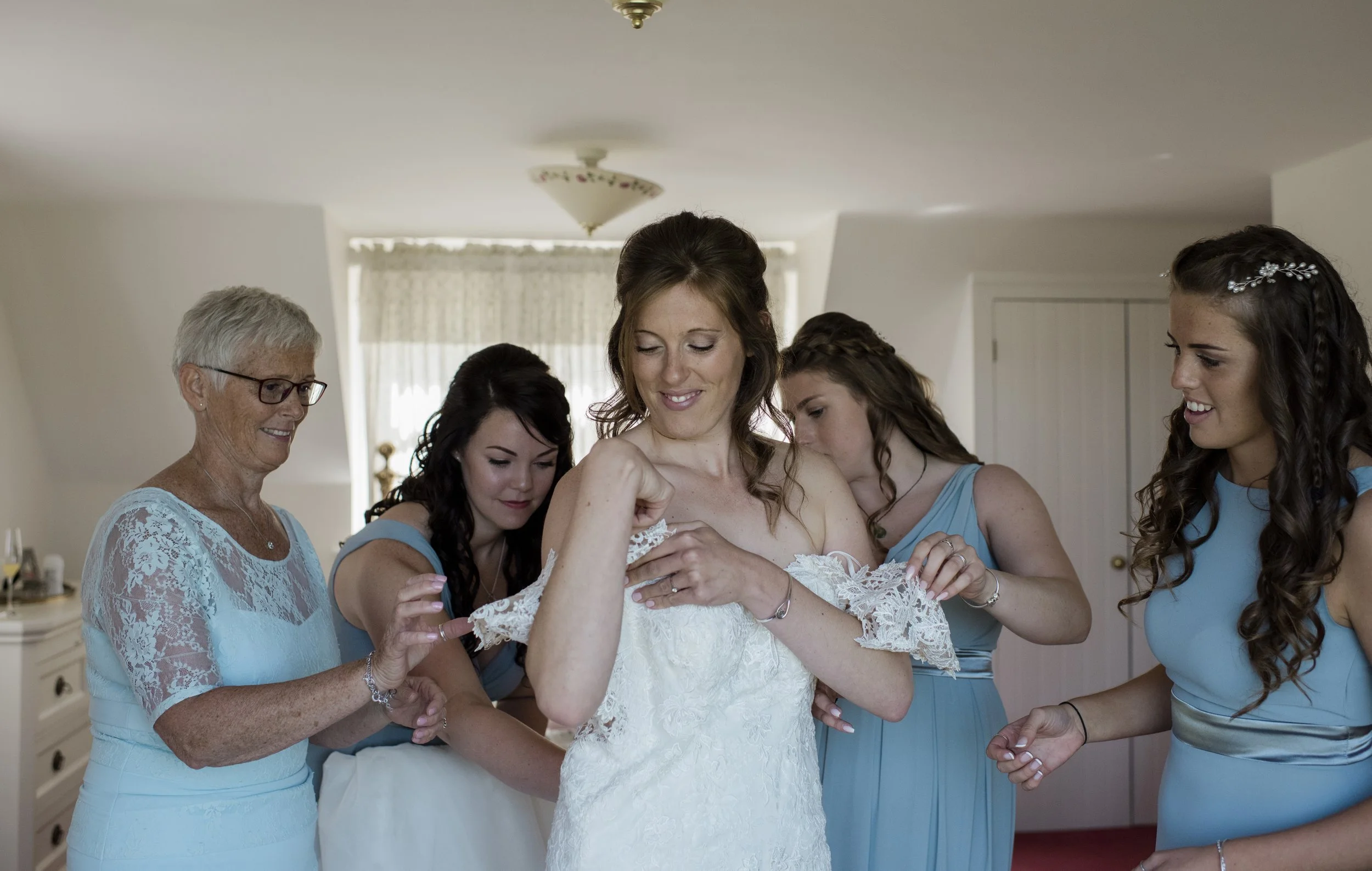 A bride with long brown hair and a lace dress is surrounded by five women, all wearing matching light blue dresses, inside a well-lit room with a ceiling light fixture and conditioned curtains. The women are helping the bride with her wedding dress.