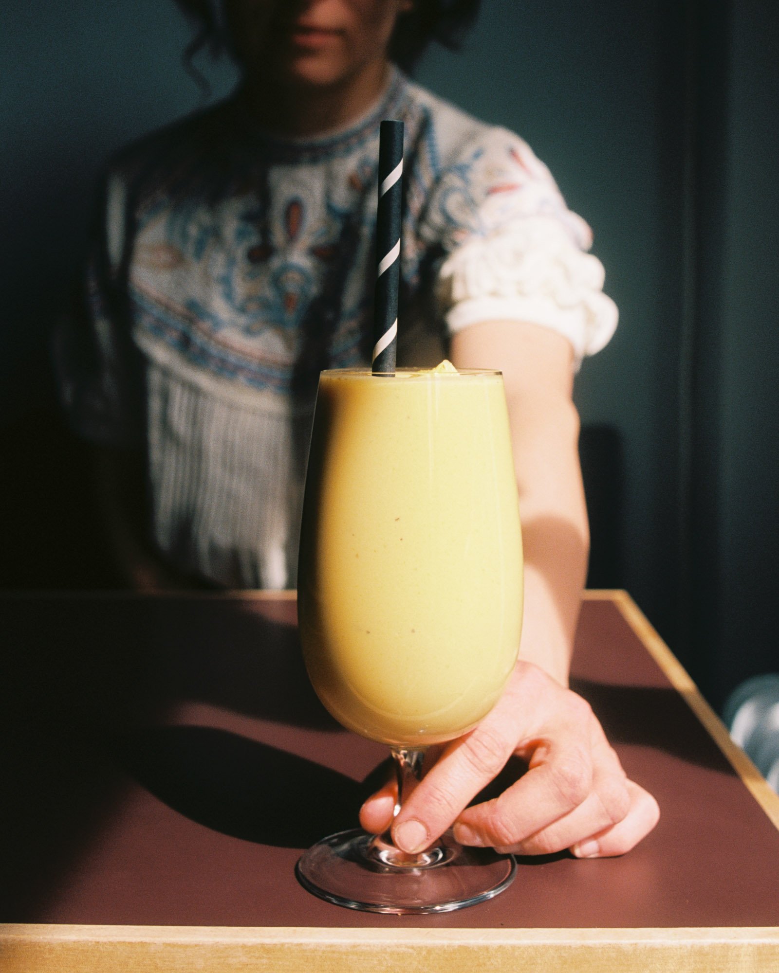 A person holding a yellow drink in a glass with a black and white striped straw.