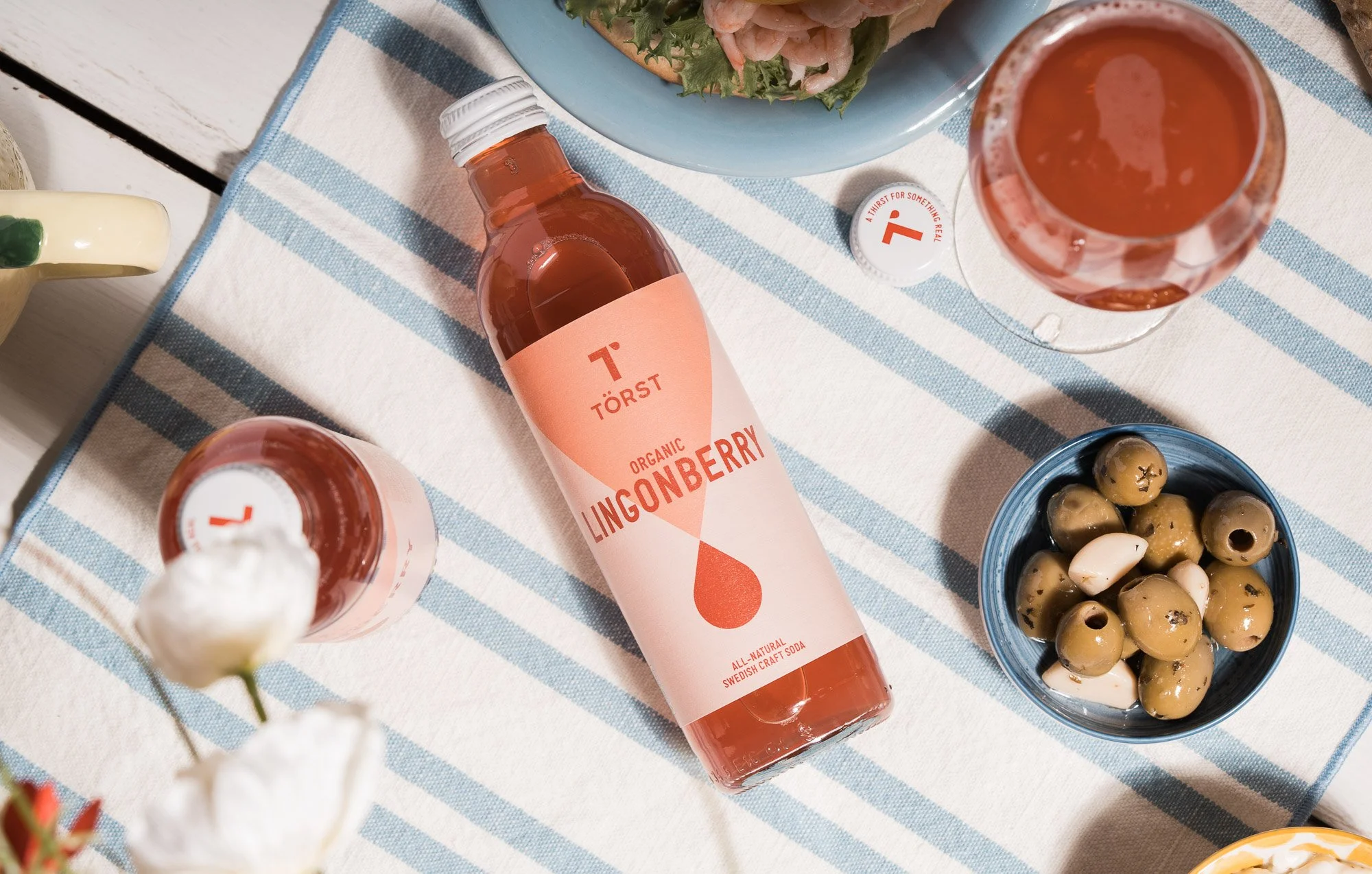 A bottle of Törst organic lingonberry soda placed on a white and blue striped tablecloth, surrounded by various food items including a bowl of green and black olives, a plate with seafood salad, a glass of soda, and a partially visible cup with a gre