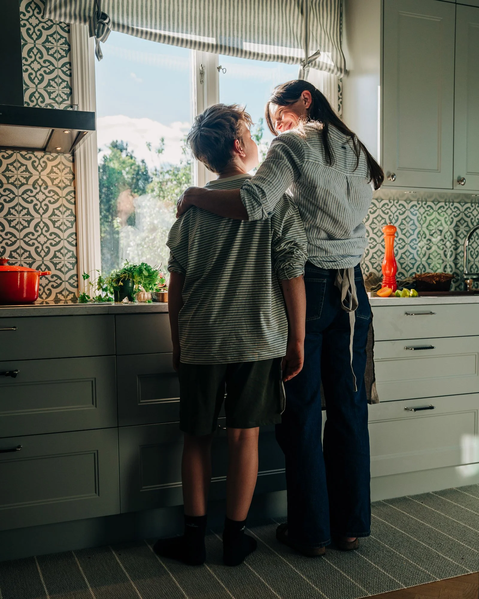 Two women sharing an intimate moment in a kitchen, standing close to each other with one woman holding the other's waist, near a window with sunlight and patterned curtains.