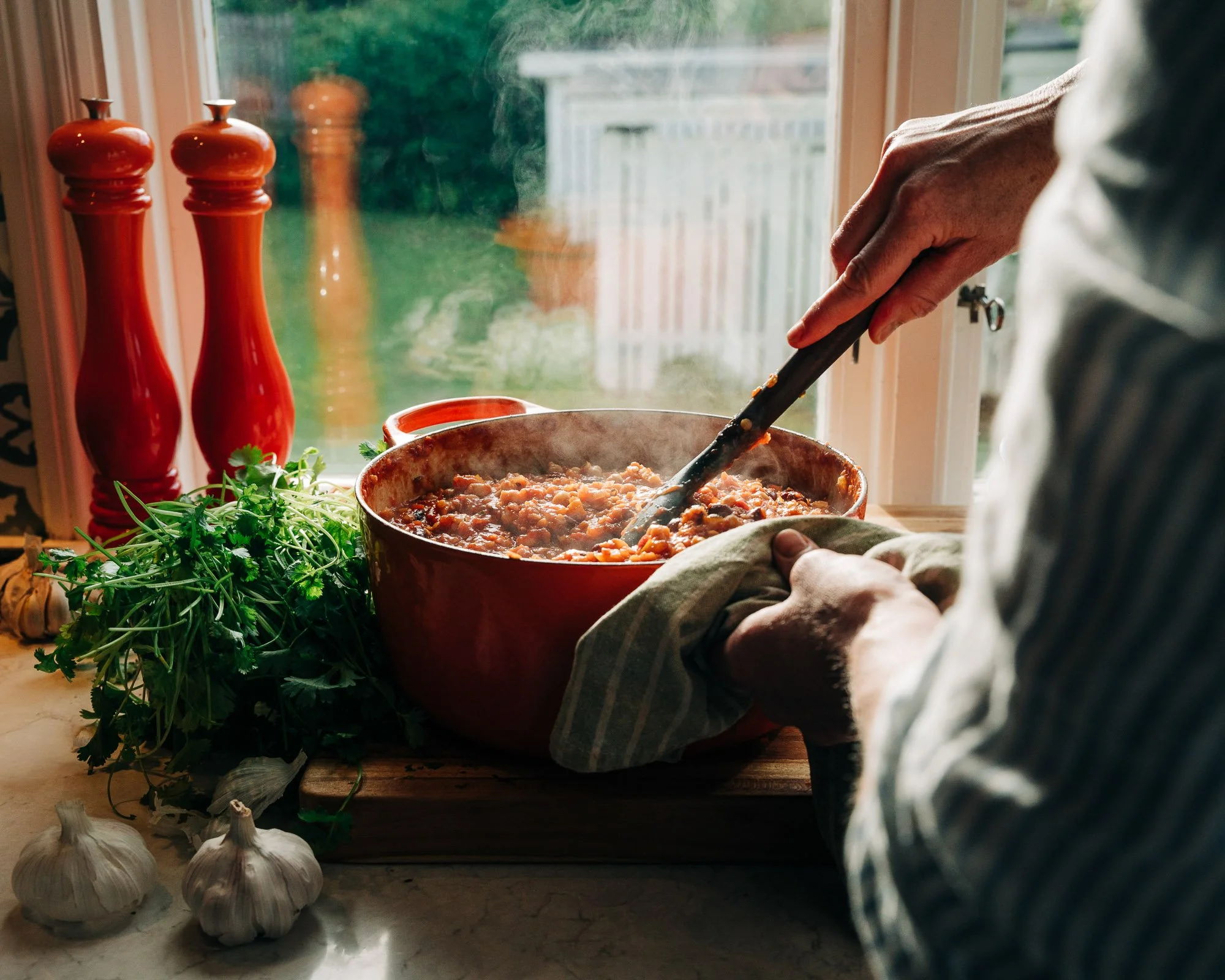 Person stirring a pot of cooked food with a spoon in a cozy kitchen near a window, with garlic bulbs and fresh herbs on the counter.