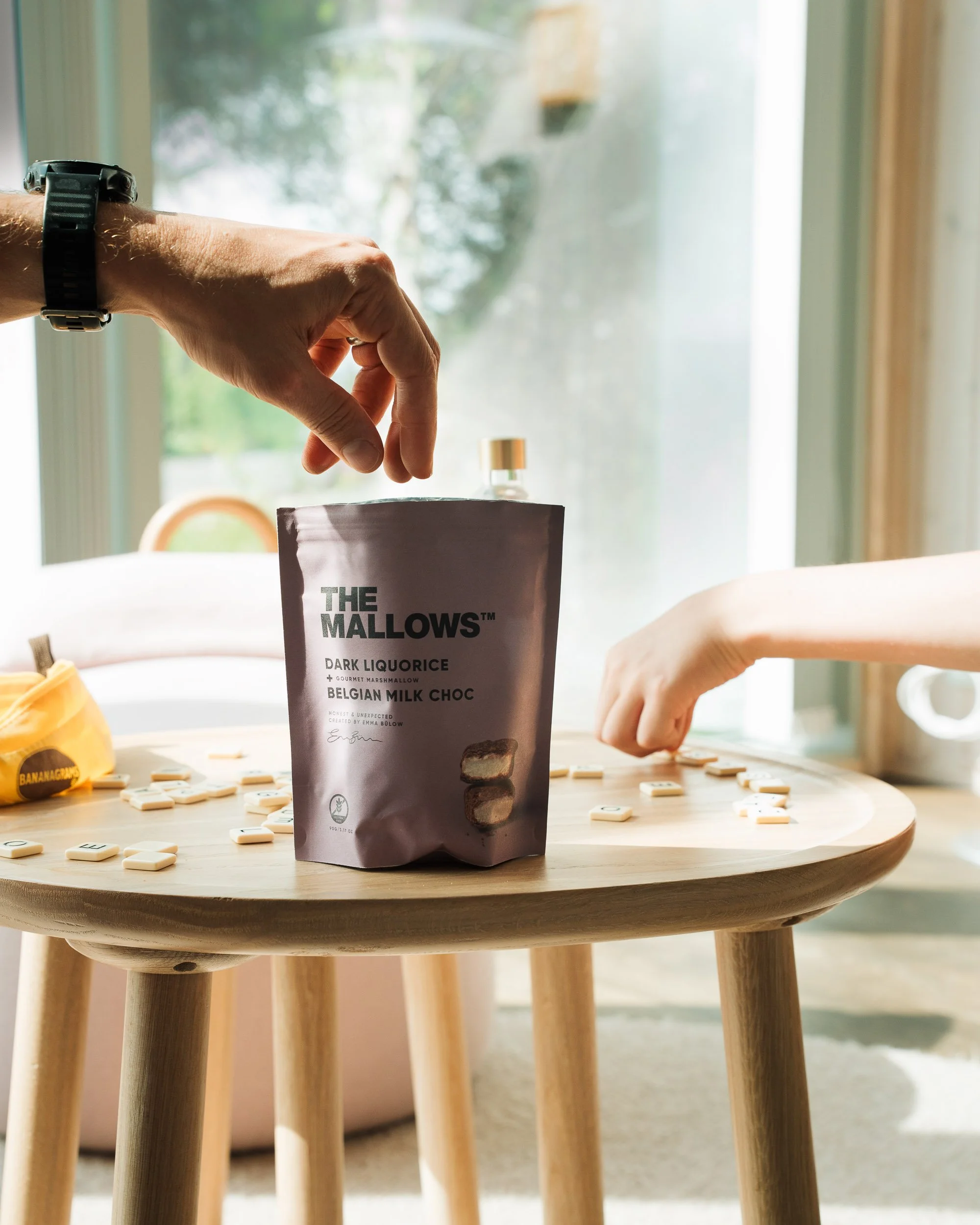 Close-up of a person's hand reaching into a bag of 'The Mallows' dark liquorice and Belgian milk choc, with scattered letter tiles on a wooden table, sunlight coming through a window in the background.