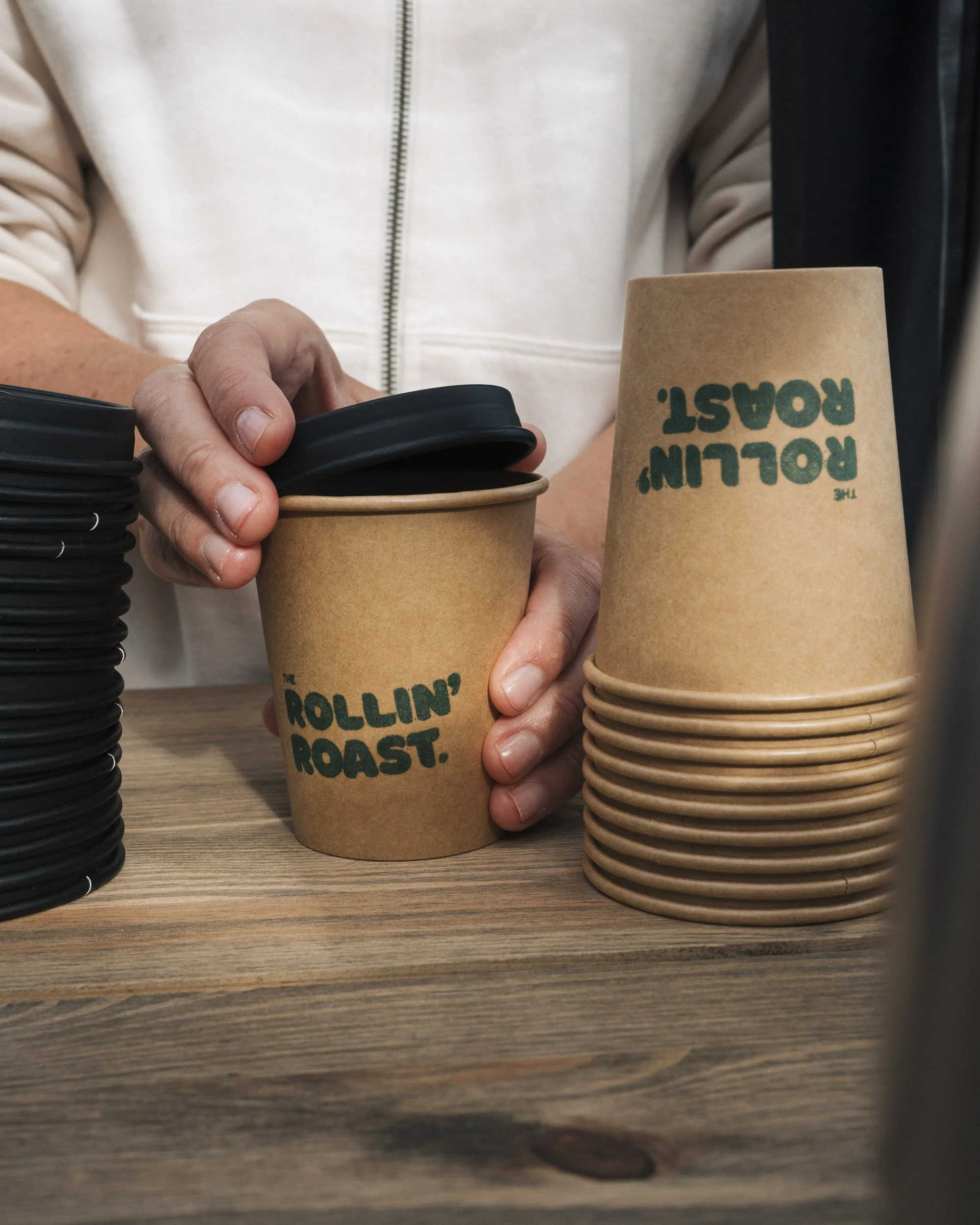 Person holding paper coffee cup with the text 'The Rollin' Roast' printed on it, stacks of similar cups, and black plastic lids on a wooden surface.