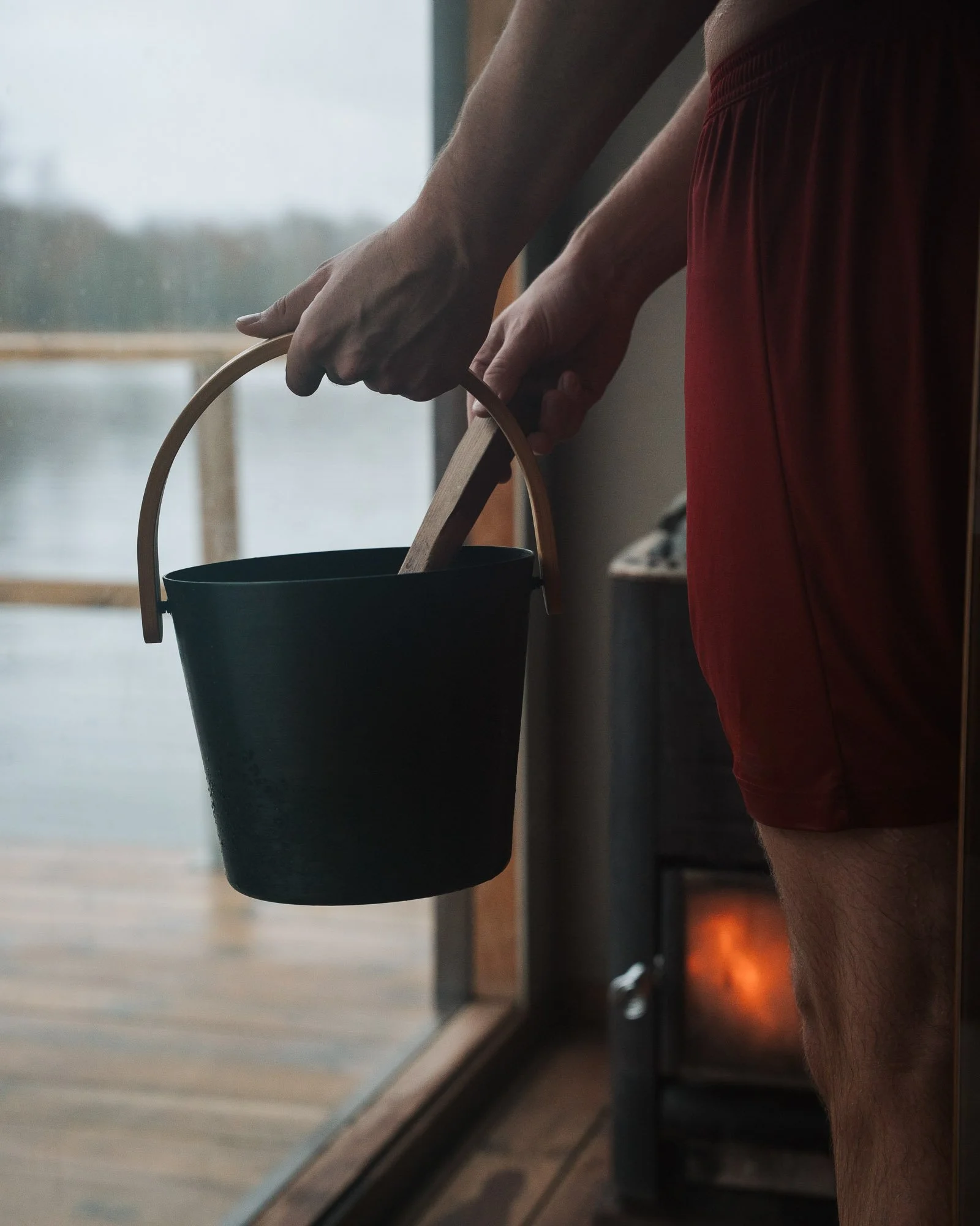 A person wearing red shorts is holding a black bucket with a handle, standing near a window with an outdoor view.