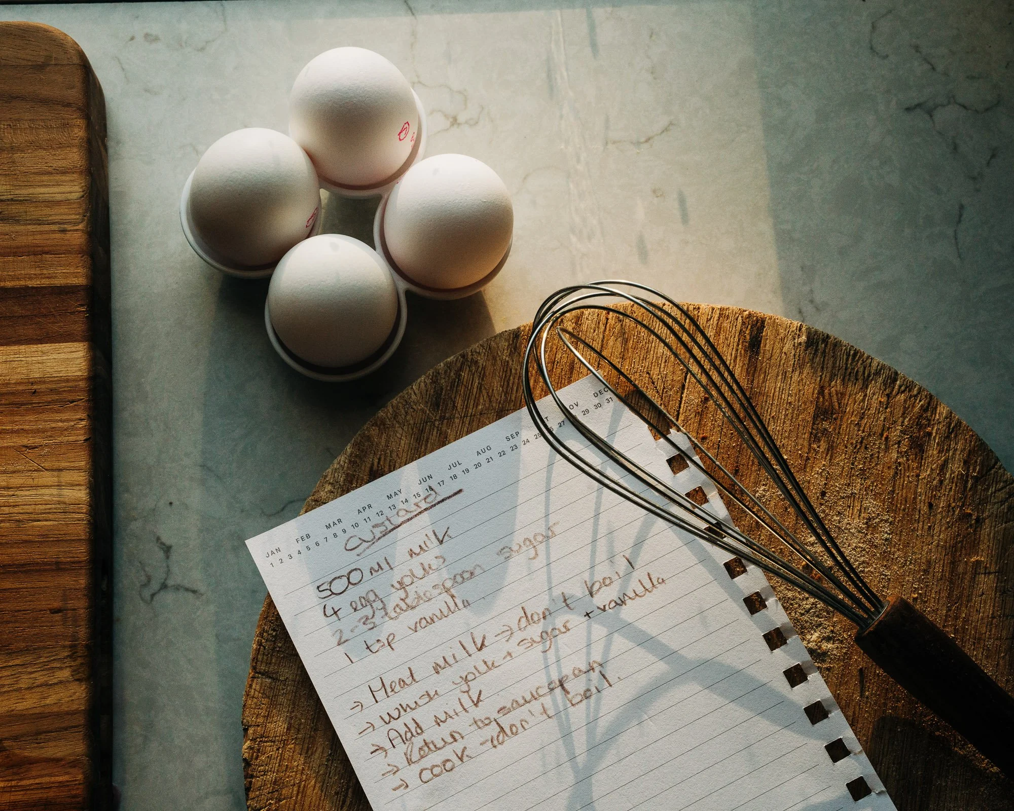 A handwritten recipe for vanilla cupcakes on lined paper, placed on a wooden cutting board with a metal whisk and a calendar page visible. Four white eggs are grouped together on a marble surface, with part of a wooden cutting board and a paper with 