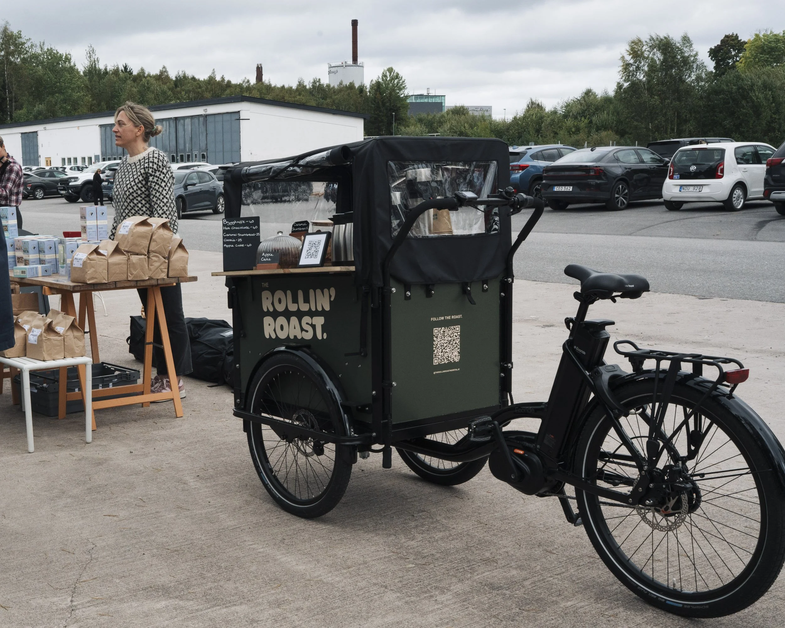 A woman at a market stall with packaged food items, a bicycle parked in front, and a food cart with a QR code and menu sign, in a parking lot with cars and trees in the background.